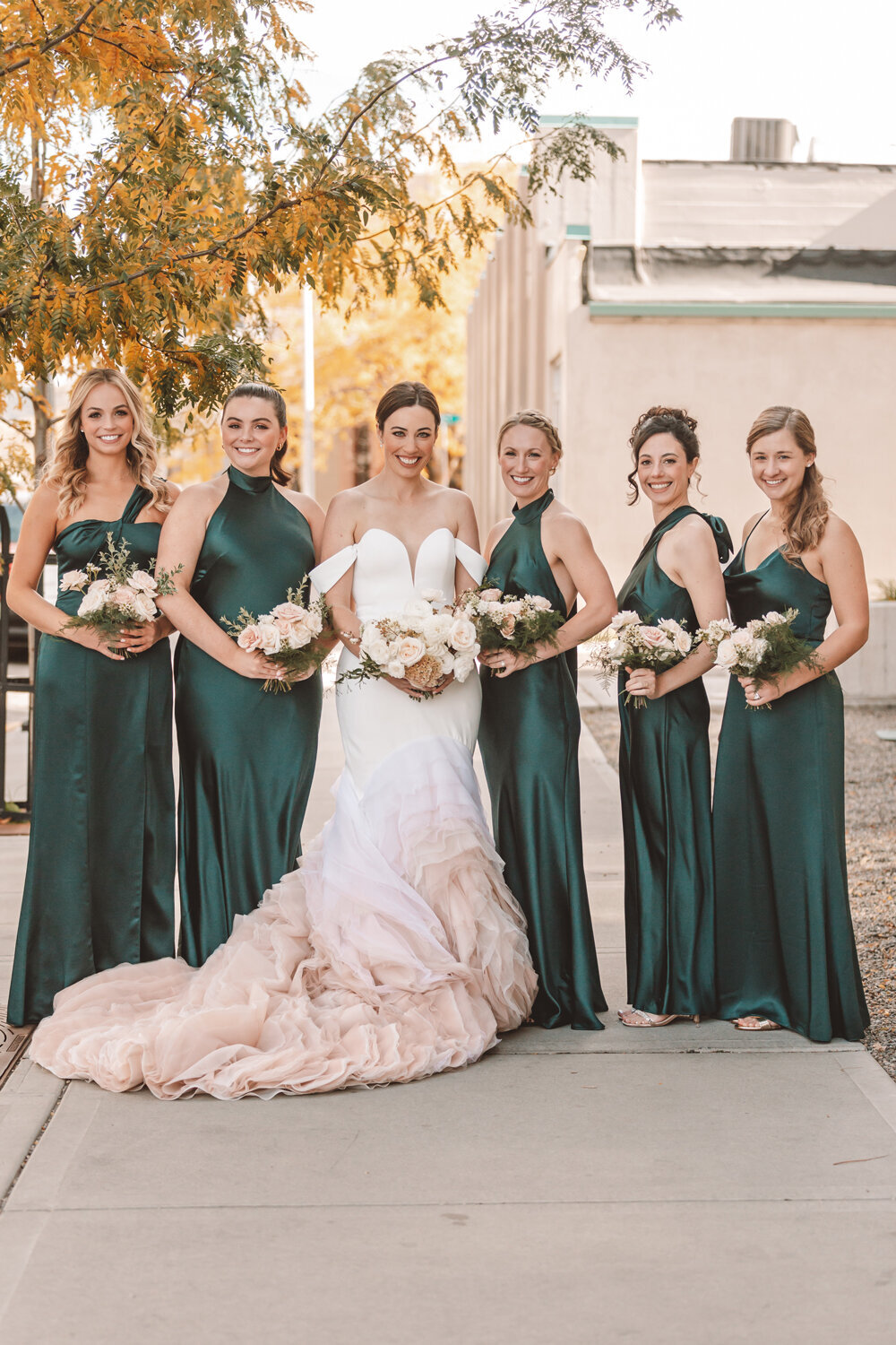 Bride and bridesmaids in emerald green satin dresses, holding blush floral bouquets, surrounded by fall foliage at a fall wedding at Kimpton Armory, Bozeman, MT.