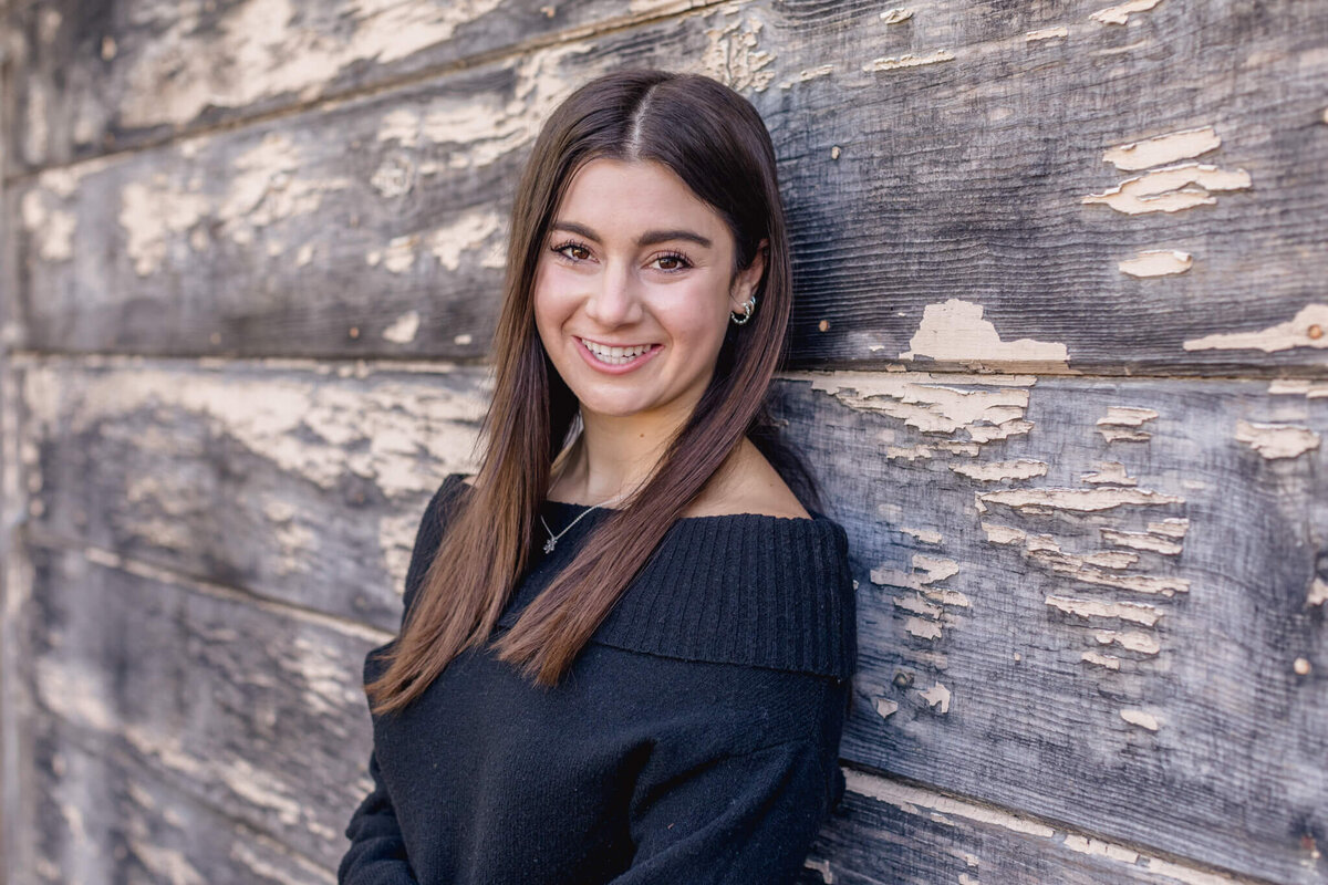 A close-up of a young woman with long, brown hair wearing a black sweater leaning her back against a grey and beige wooden wall, looking sidelong at the camera and smiling near Katie Wheeler Library in Irvine.