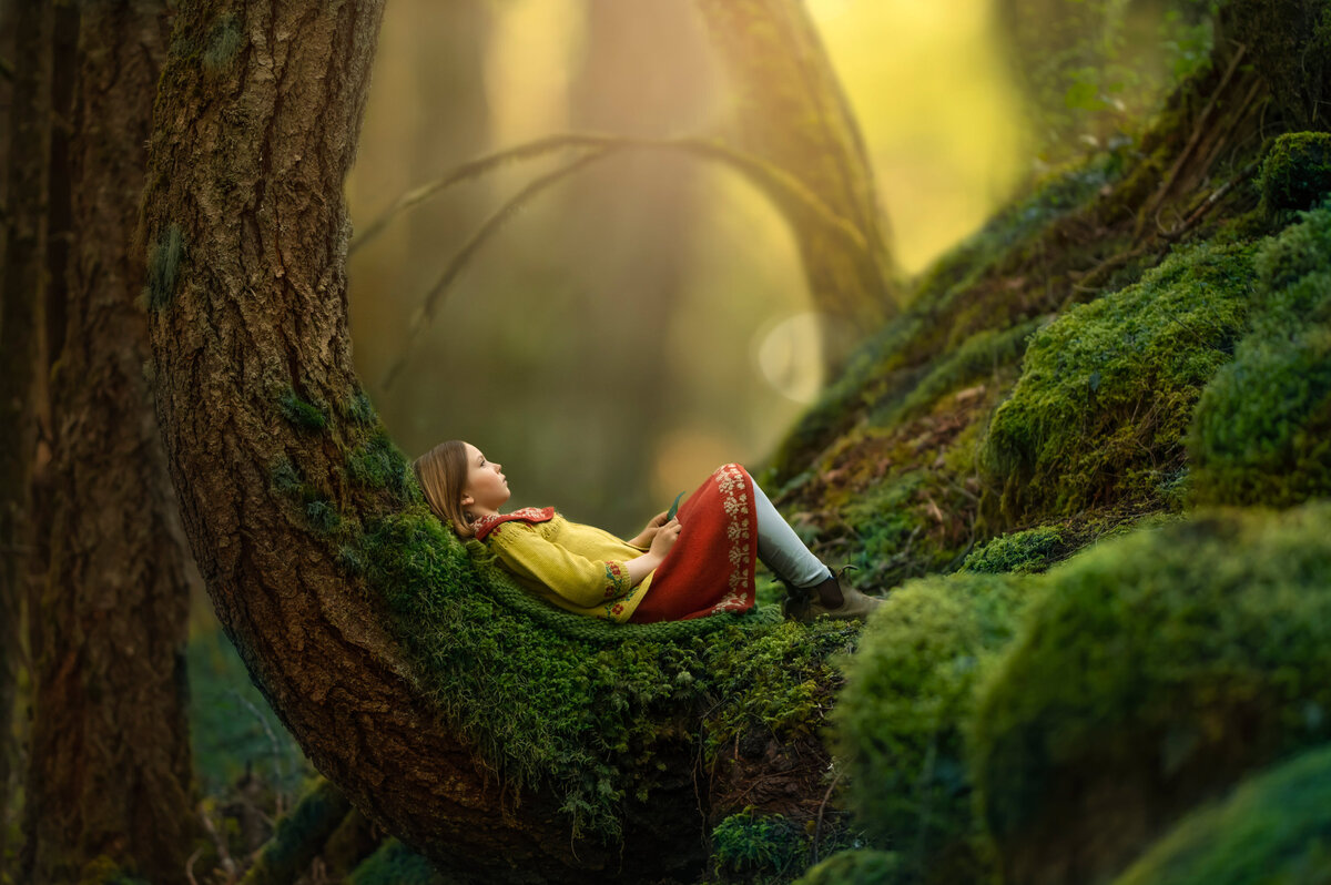 A girl rests in bend of a moss-covered tree during her storybook-style enchanted forest photo shoot in Waukesha, WI.