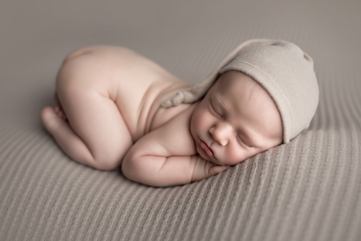 Naked newborn baby wearing a soft taupe bonnet, posed in the curled-up sleeping position on a neutral taupe blanket, captured by DFW newborn photographer.