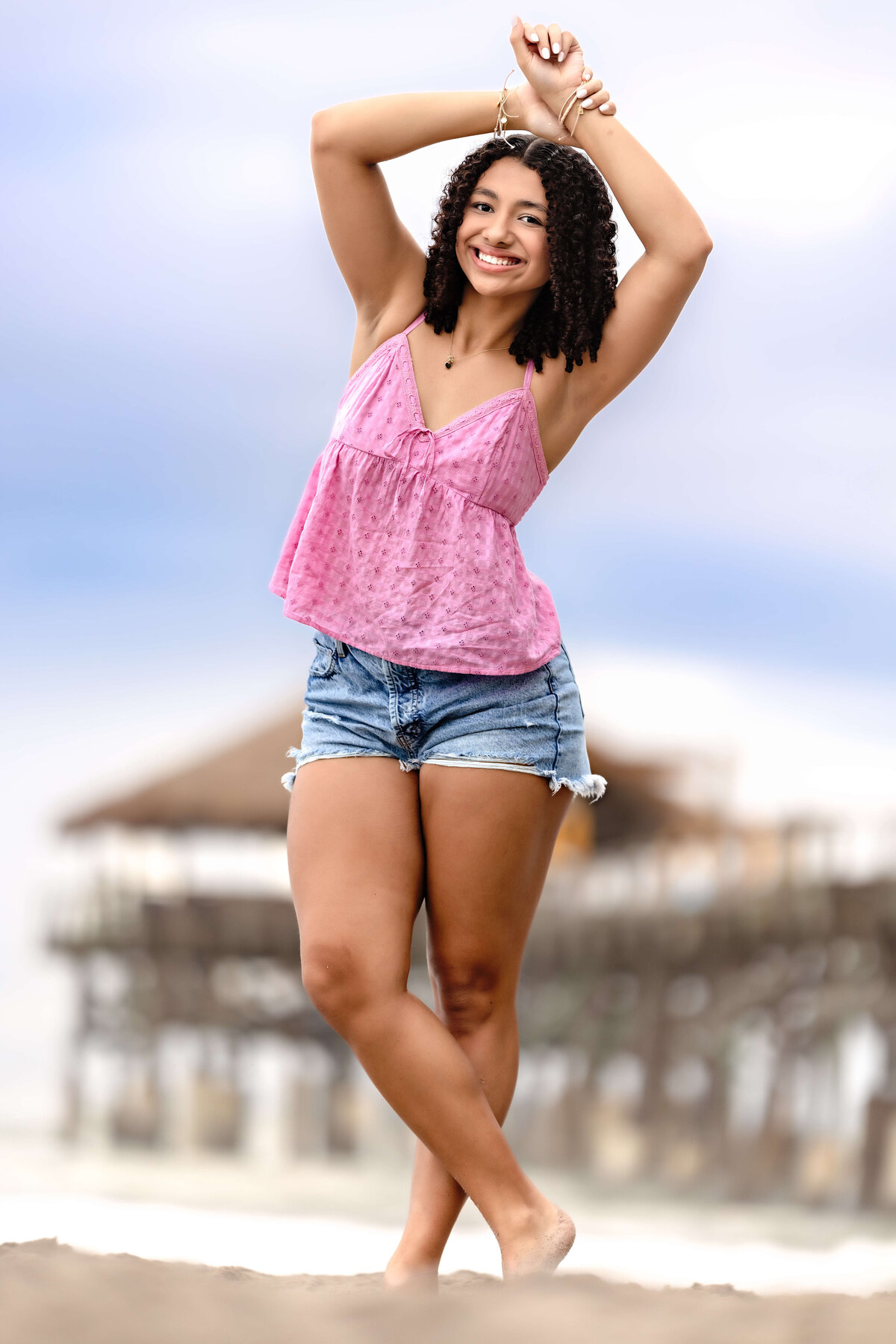 Fun, Gen Z-inspired senior portrait with a girl in jean shorts and a pink top, hands above her head, stormy sky and Cocoa Beach Pier behind her.