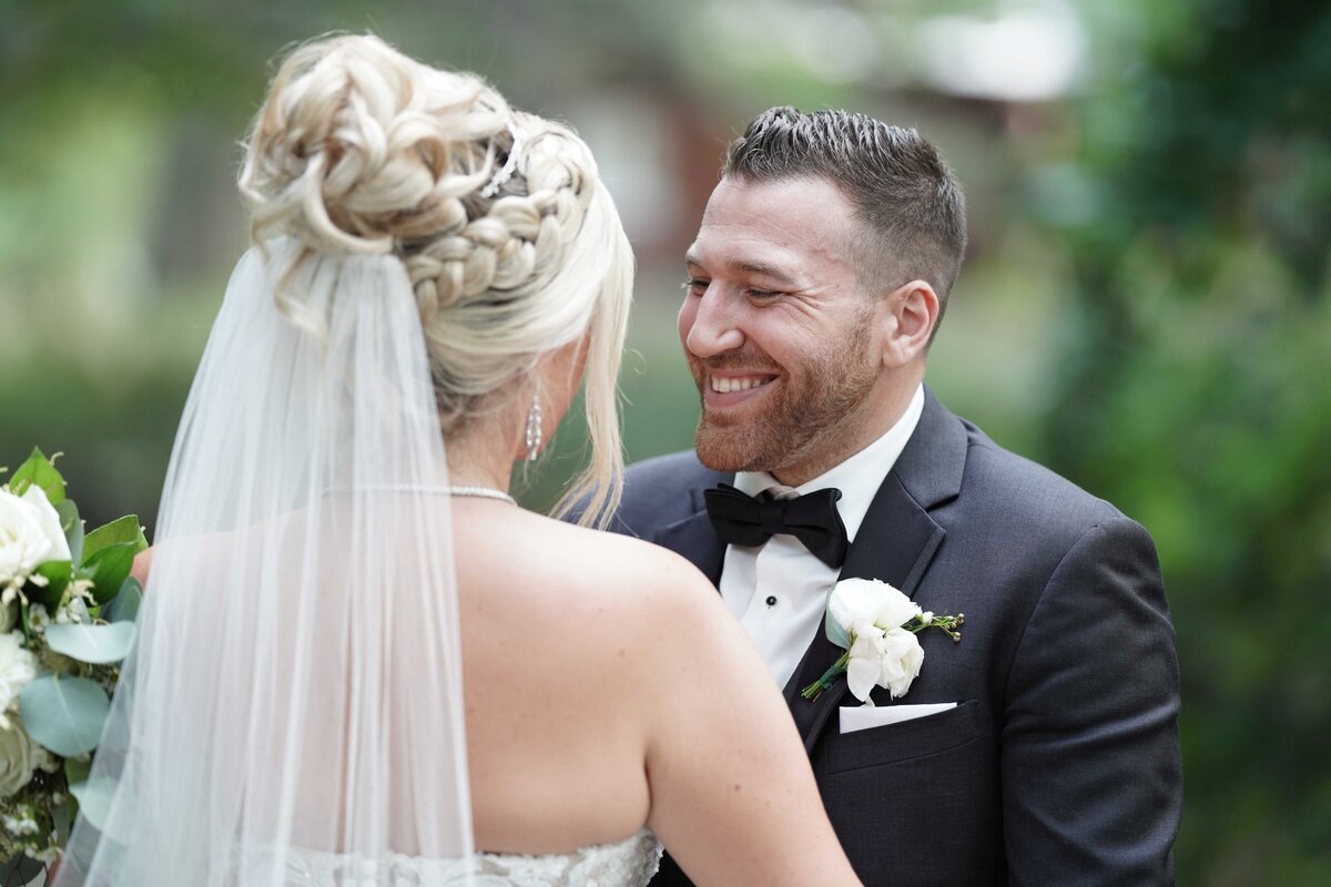 A close-up shot of the groom smiling warmly at his bride, showcasing their deep connection and joy. The image captures the groom’s genuine expression and the loving gaze he directs towards his bride, highlighting a moment of affection and happiness during their wedding.