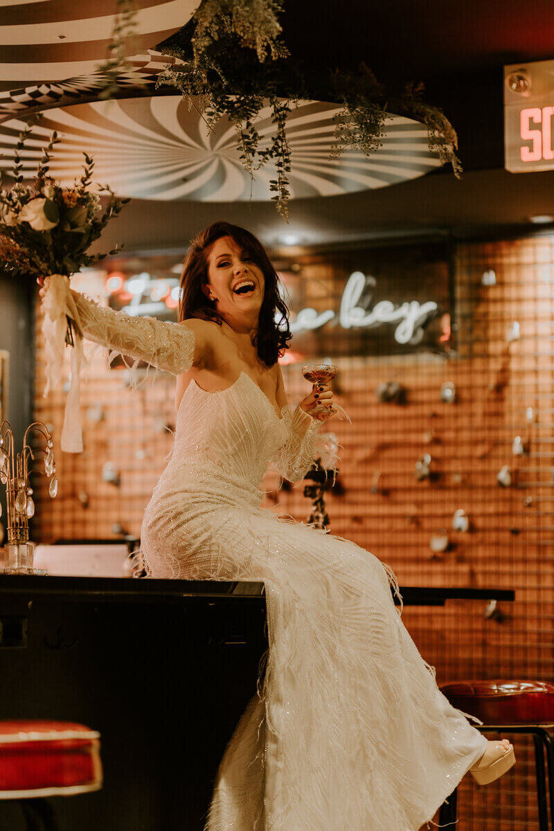 Mariée en robe blanche vintage avec longs gants brandissant son bouquet, assise sur un bar dans un décor vintage aux tons chauds lors d'une séance photo mariage.