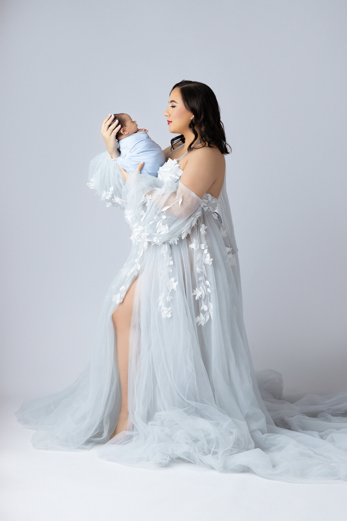 Beautiful mom in flowing sheer gown holding her swaddled baby, photographed by a DFW newborn photography studio.