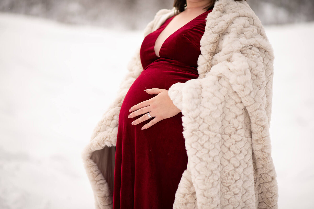 An up close image of a baby belly. The woman wears a red dress and fluffy beige blanket. She has one hand on her belly. Portrait by Brittany Danielle
