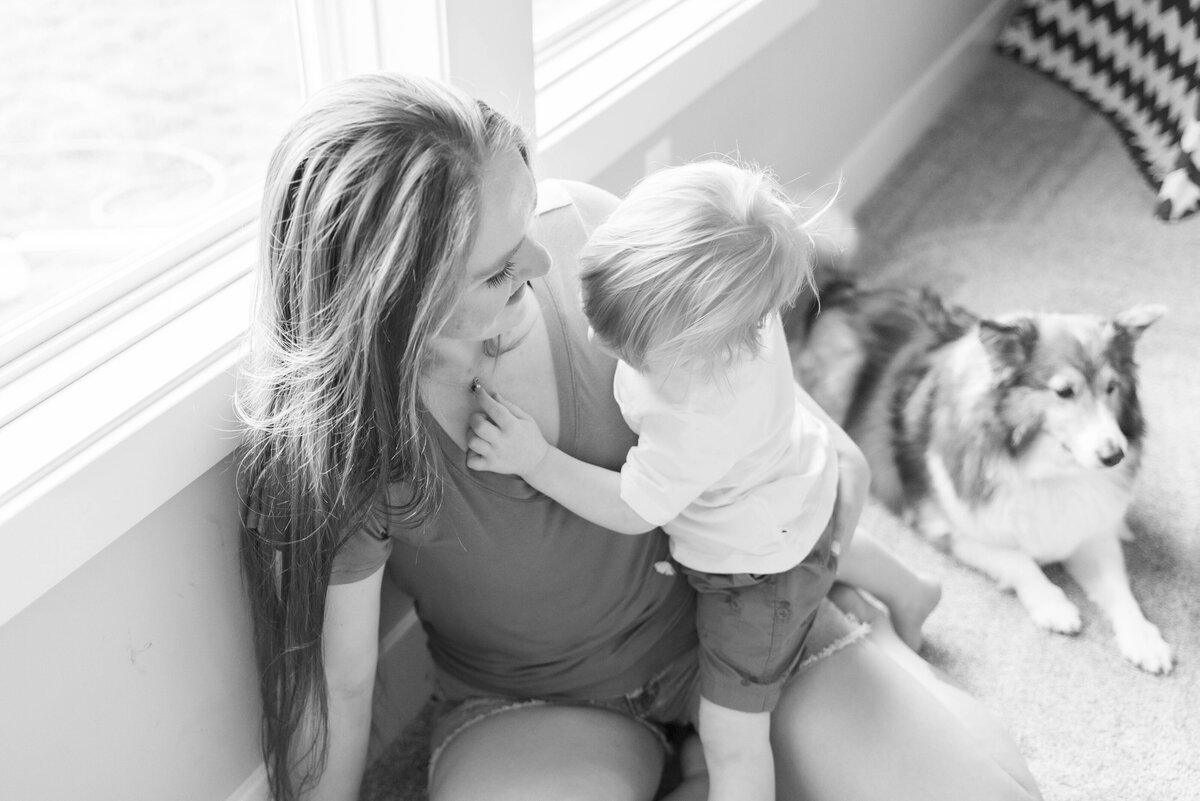 Black and white photo of a mother holding her toddler by a window with their dog nearby during a cozy in-home family session.