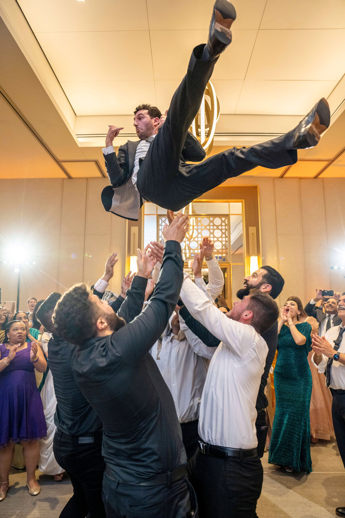 A group of people in formal attire toss a man in a suit into the air at a lively indoor celebration. The crowd around them watches with excitement, some clapping and capturing the moment on their phones. The room is brightly lit and decorated elegantly.