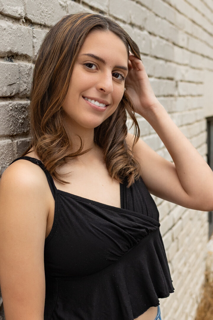 A senior girl leaning against a white brick wall looking at the camera and smiling in Lawrence, KS