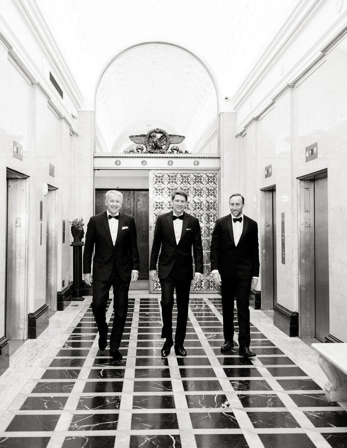 The photo was taken at Washington National Cathedral in Washington D.C. by photographer Twah Dougherty. The three men in tuxedos were captured elegantly walking down a grand hallway with marble flooring and ornate decor. The setting appears to be an upscale, classical building, possibly a hotel or a historic venue. Large decorative elevators flank either side of them, enhancing the luxurious atmosphere depicted in the black and white image. As for current films as videographers, I don't have that specific information, but I can certainly assist you with any other queries you may have. How else can I help you today?