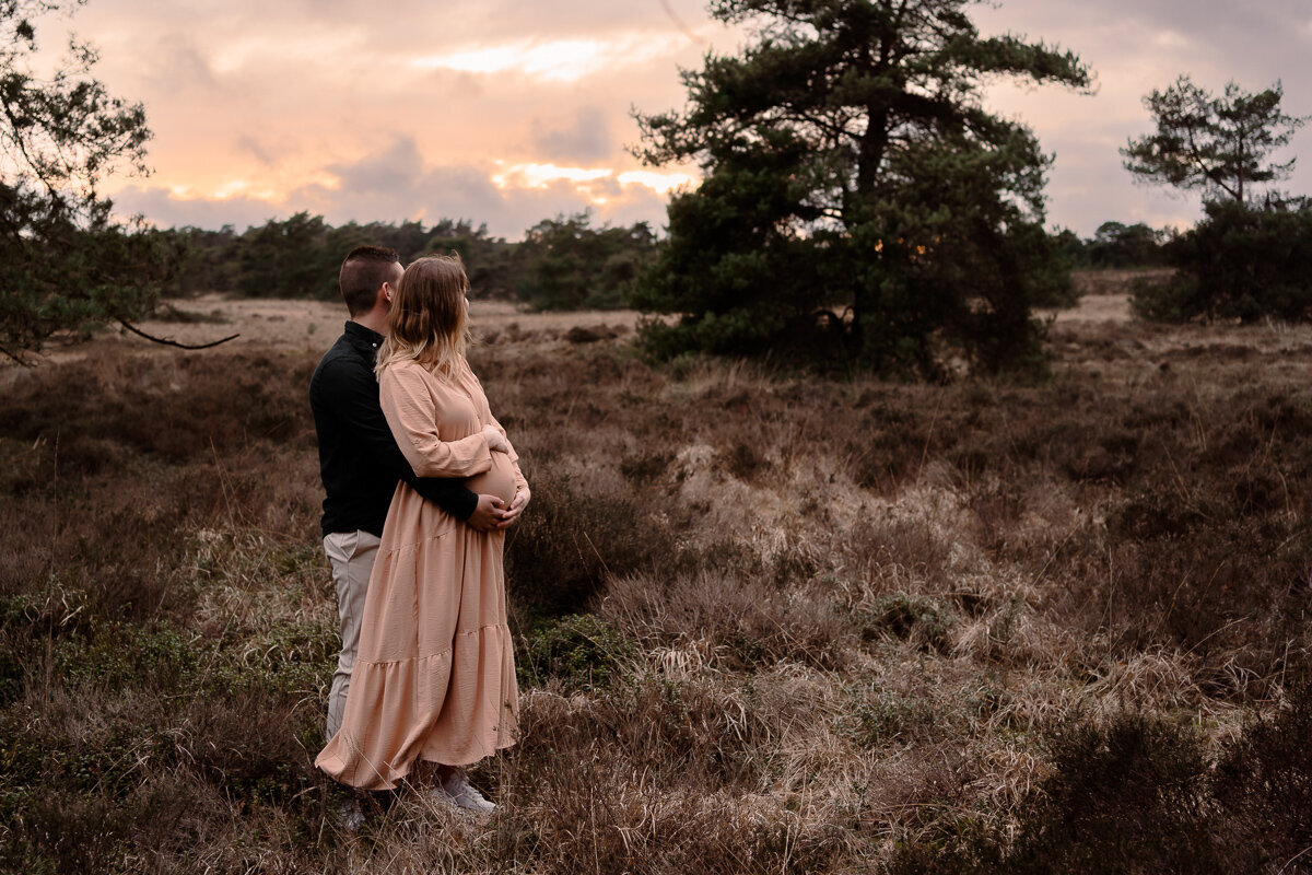 Zwangere vrouw staat tegen haar partner aan terwijl zij beiden hun handen op haar buik hebben
