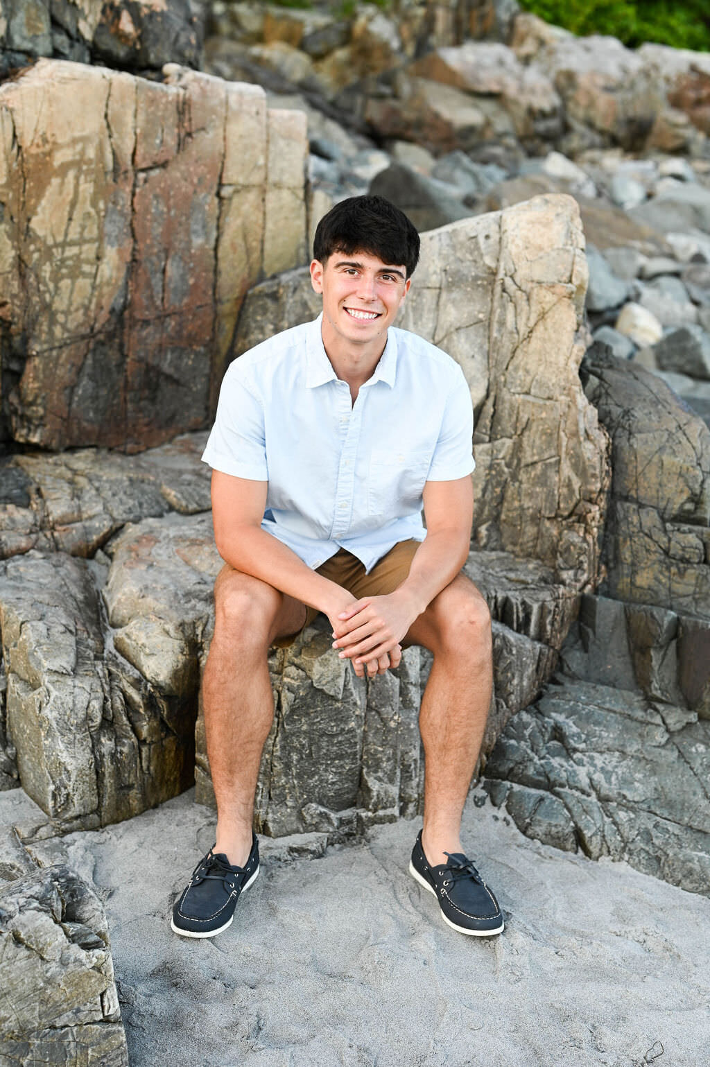 A boy smiling and sitting on rocks in the sand.