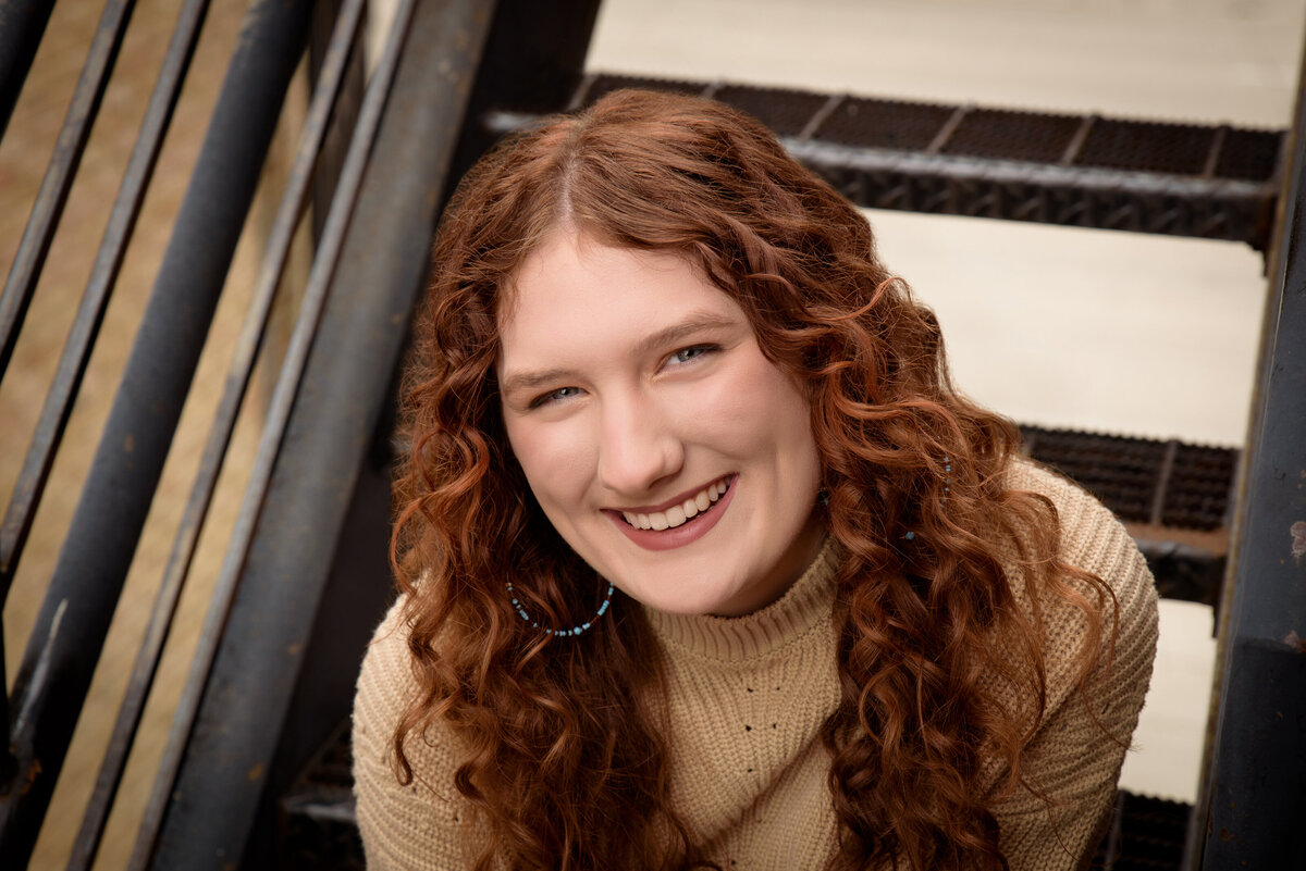 Green Bay East High School senior girl sitting on metal stairs wearing a tan sweater and jeans in urban setting in downtown Green Bay, Wisconsin.