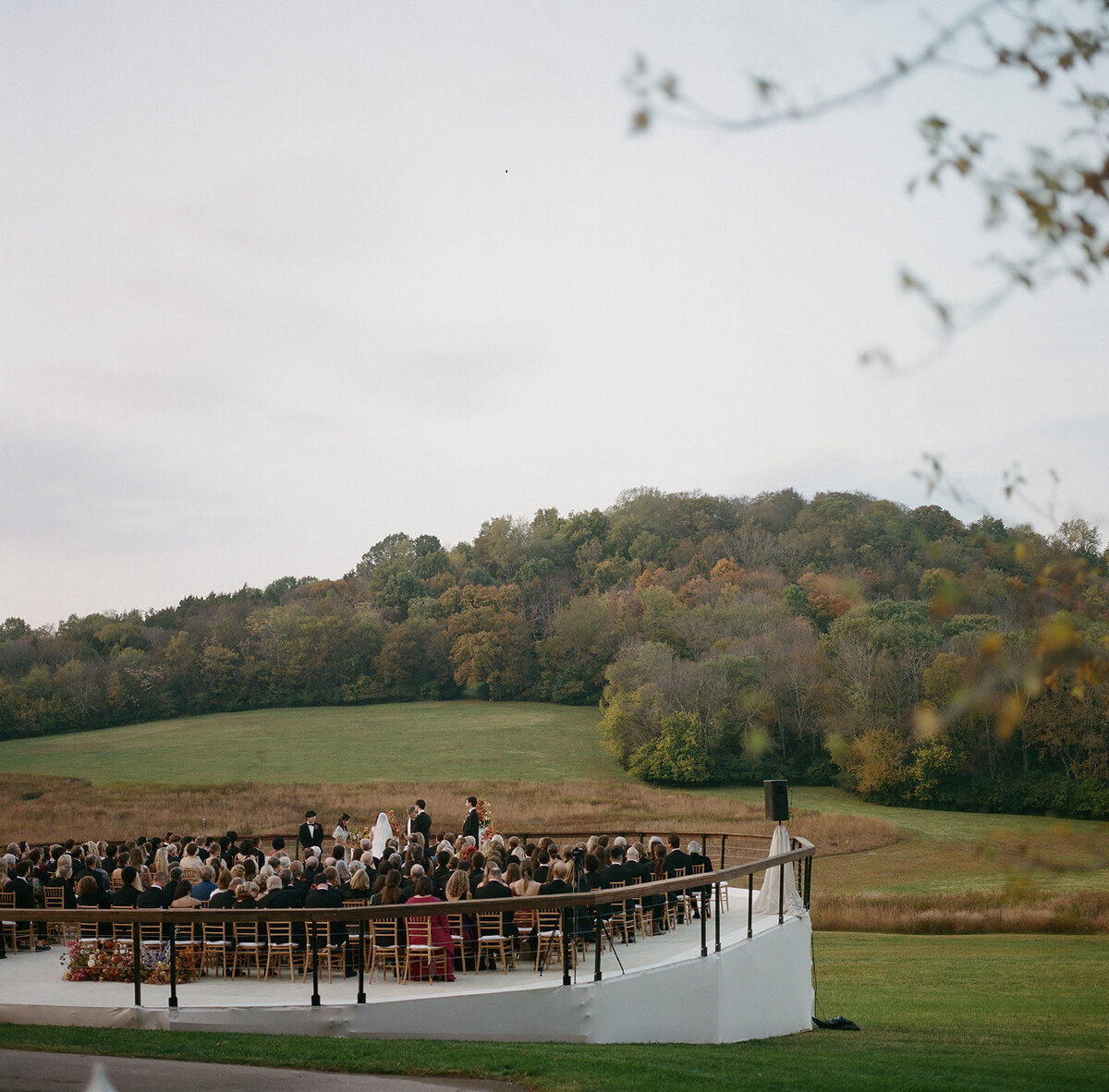 Sculptural columns wedding ceremony backdrop. Stair step white ceremony columns adorned with sculptural floral meadows in fall hues. Clear top tent build out for fall wedding reception in Tennessee hills. Wire threaded orchids, bittersweet berries, butterfly ranunculus, and tulips are highlighted in this wedding ceremony backdrop. Artistic fall color palette in copper, gold, ruby, lavender, and rust. Design by Rosemary & Finch Floral Design in Nashville, TN.