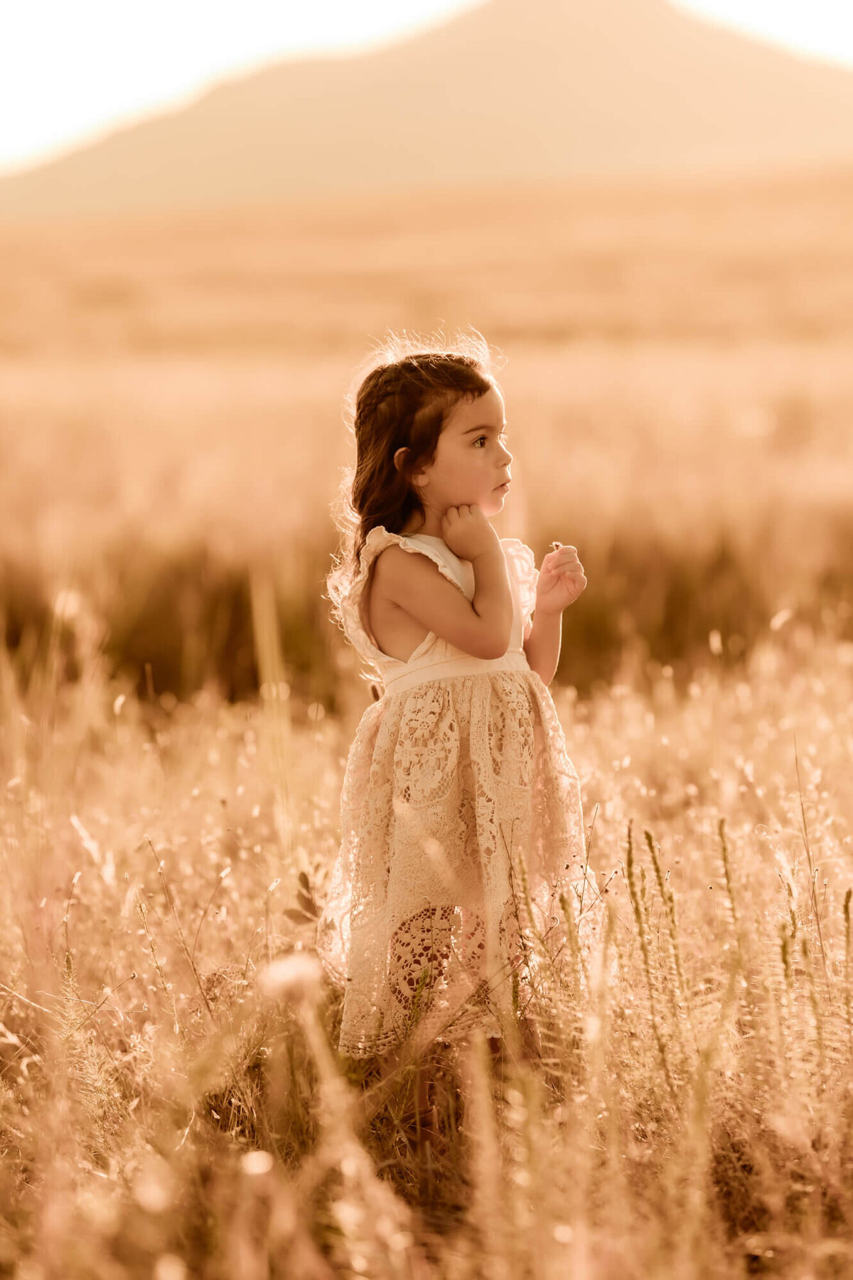 Little girl wearing a golden dress rests her hand under her chin as she looks off into the distance.