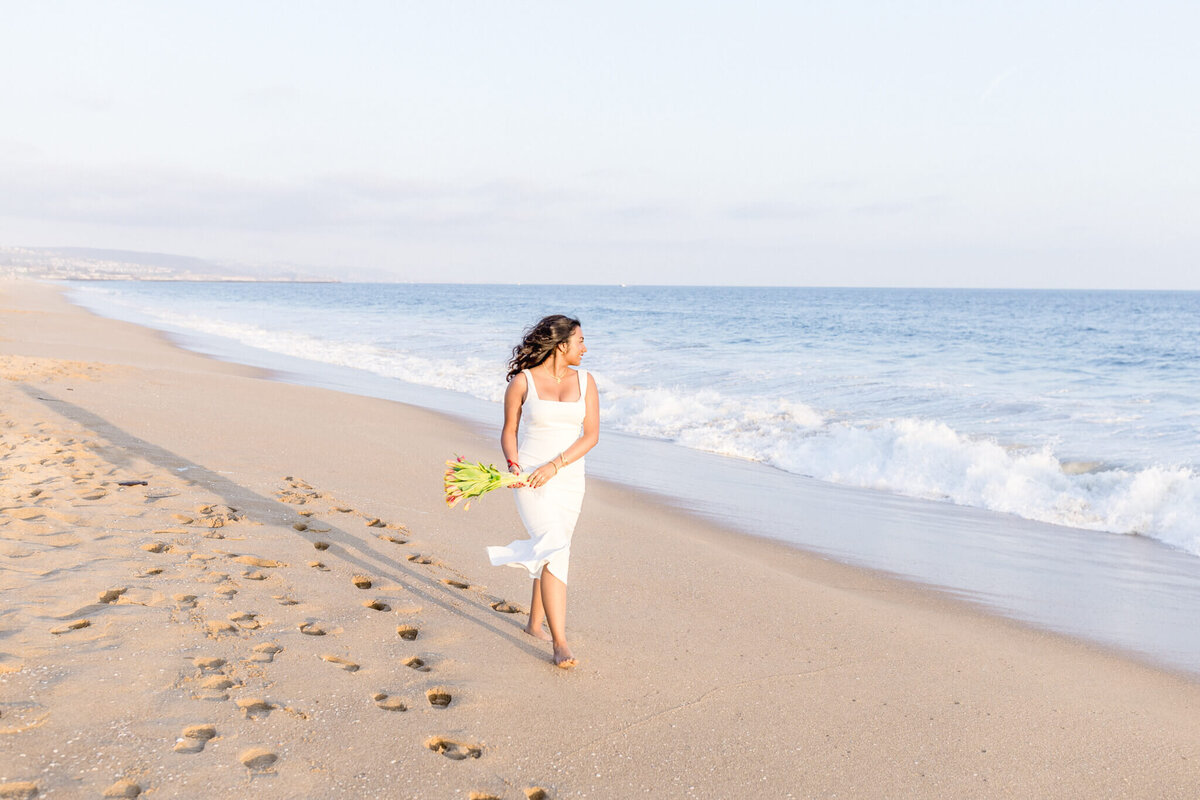 A woman in a white dress walks barefoot along a sandy beach, holding a bouquet of flowers. The ocean waves gently lap the shore under a clear sky.