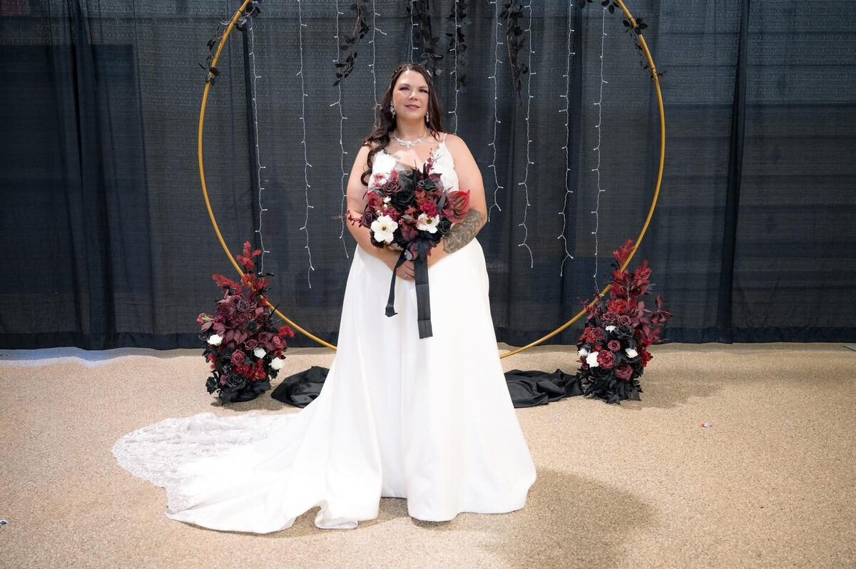 Bride in a white gown holding a dark floral bouquet standing beneath a circular arch.