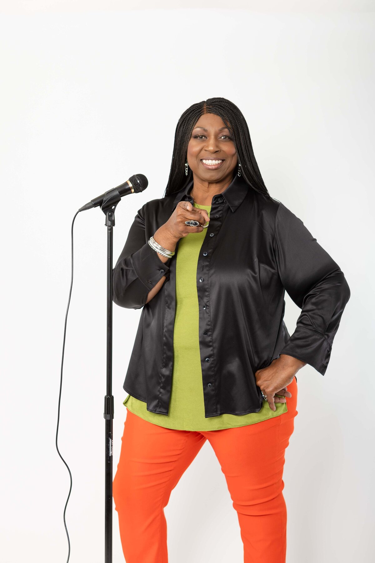 A bold and stylish woman smiles while pointing toward the camera next to a standing microphone, dressed in bright orange pants and black blouse—captured as a powerful speaker stand-up comedian brand photo.