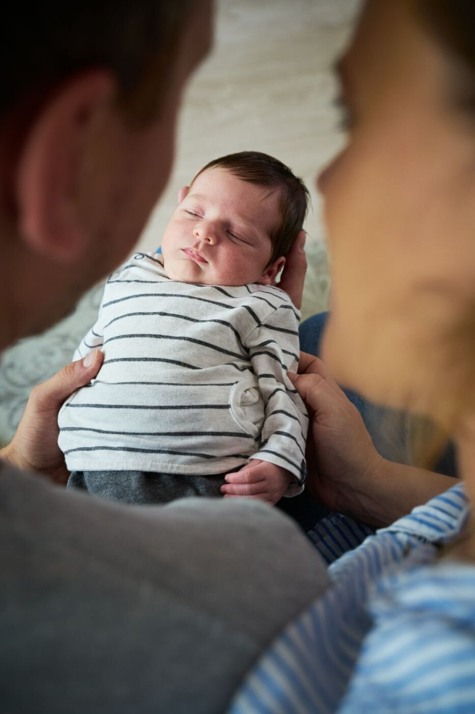  A close-up, candid photo of a newborn baby sleeping peacefully while being held by its parents. The baby is in sharp focus, wearing a striped onesie. The parents' heads and hands are in the foreground, blurred and out of focus, looking down at the baby with love.