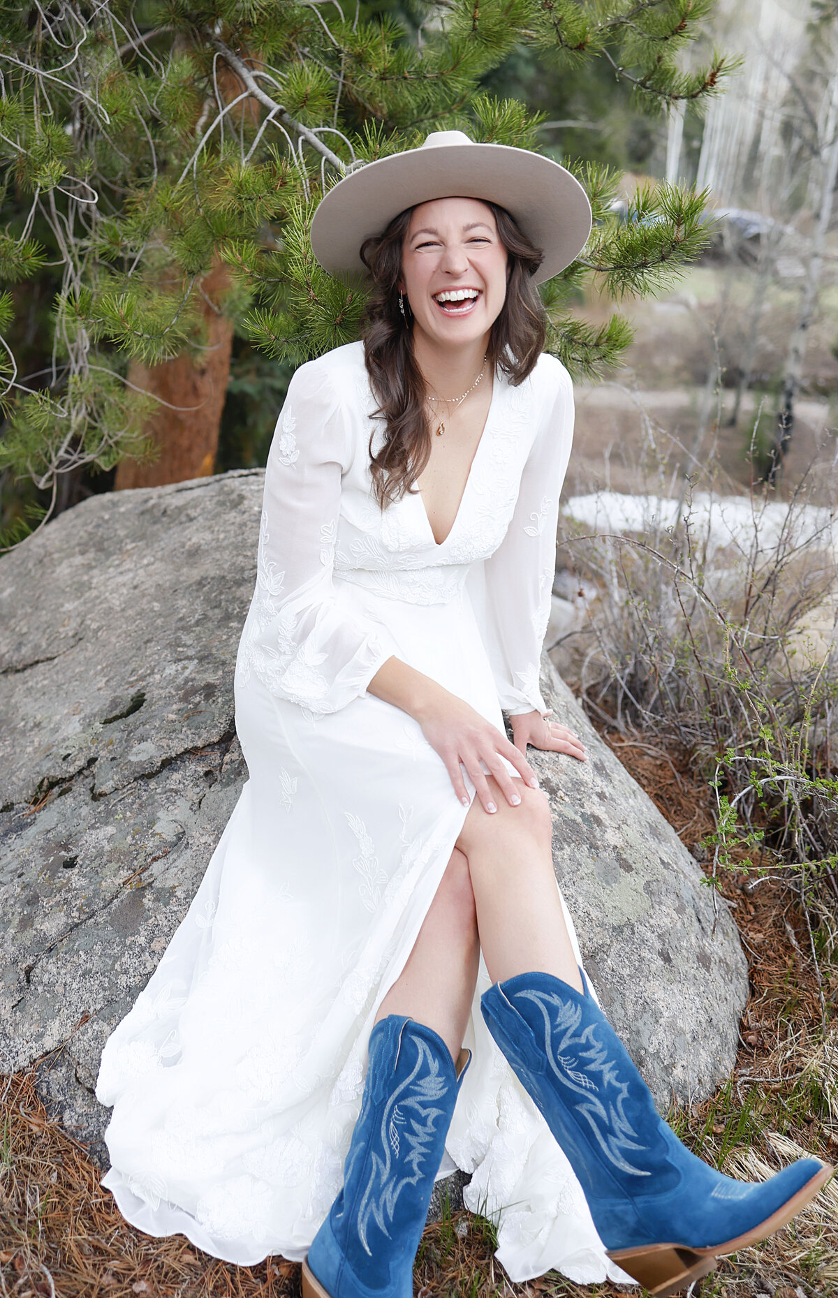 A bride laughs as she sits on a rock, showcasing her adorable blue cowboy boots and smile.