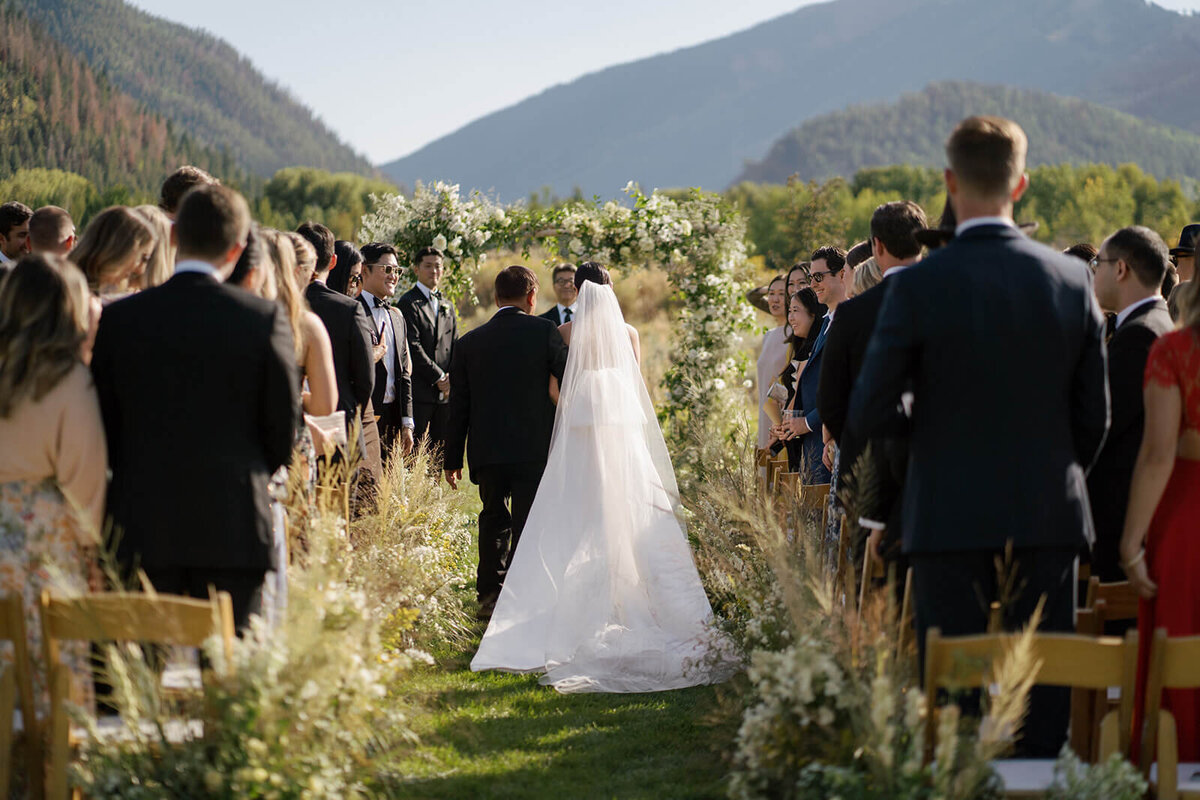 The father of the bride walks his daughter down the aisle amid beautiful Colorado mountains at her Aspen Meadows Resort wedding ceremony.