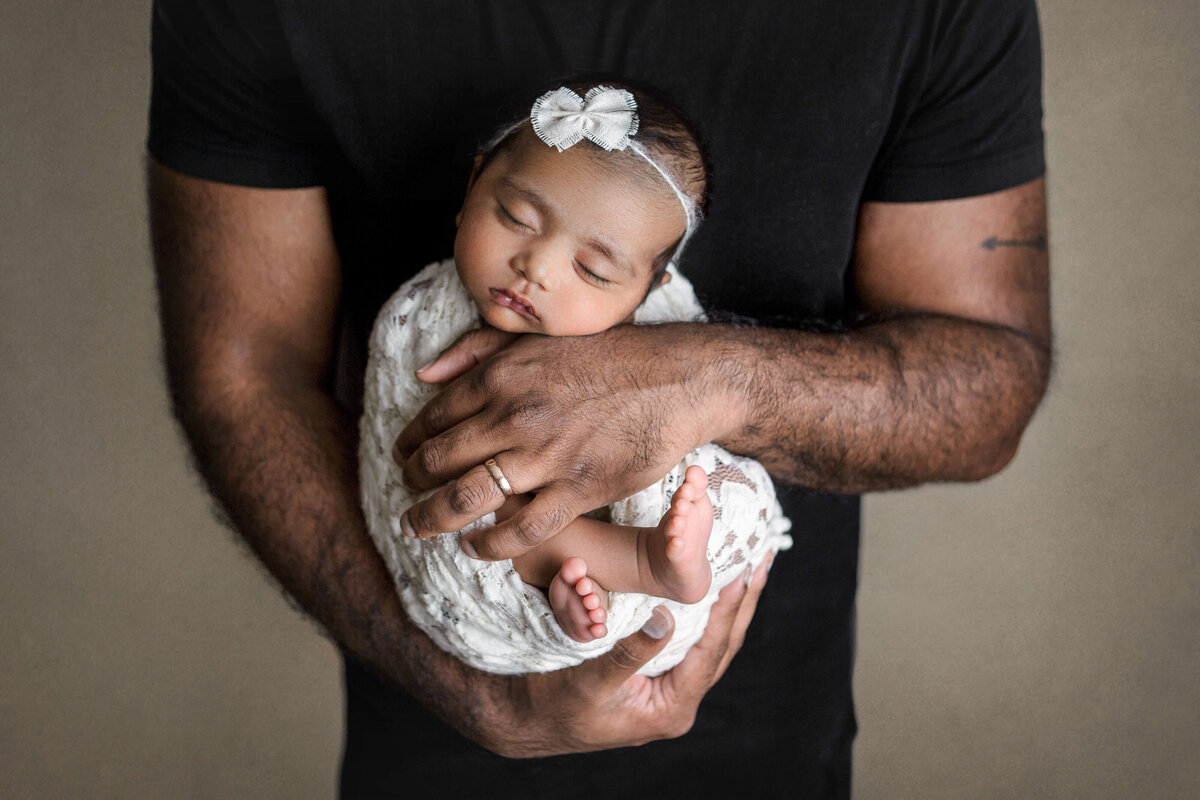 dad wearing a black shirt with his arms holding his newborn on his chest in a white lace wrap and bow headband