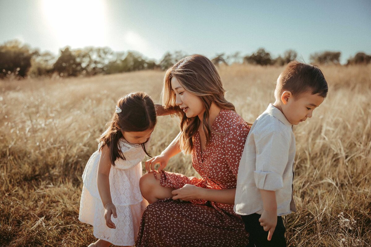 A woman is kneeling on the grass wearing a red dress. She is looking at her daughter who is wearing a white sleeveless dress. Her son is facing the other way and looking at the yellow Texas grass.