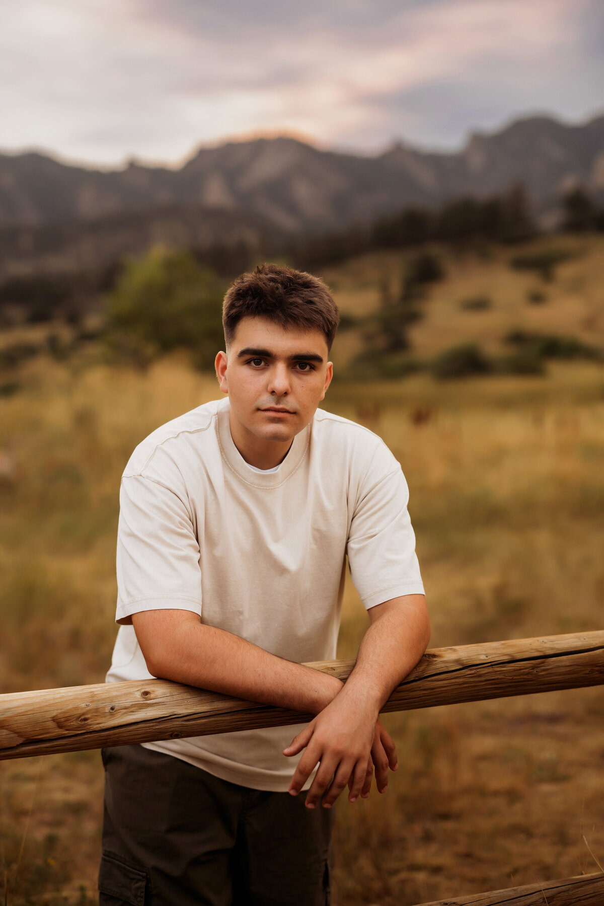 high school senior boy poses with mountains behind him and leaning over a fence for his portrait session