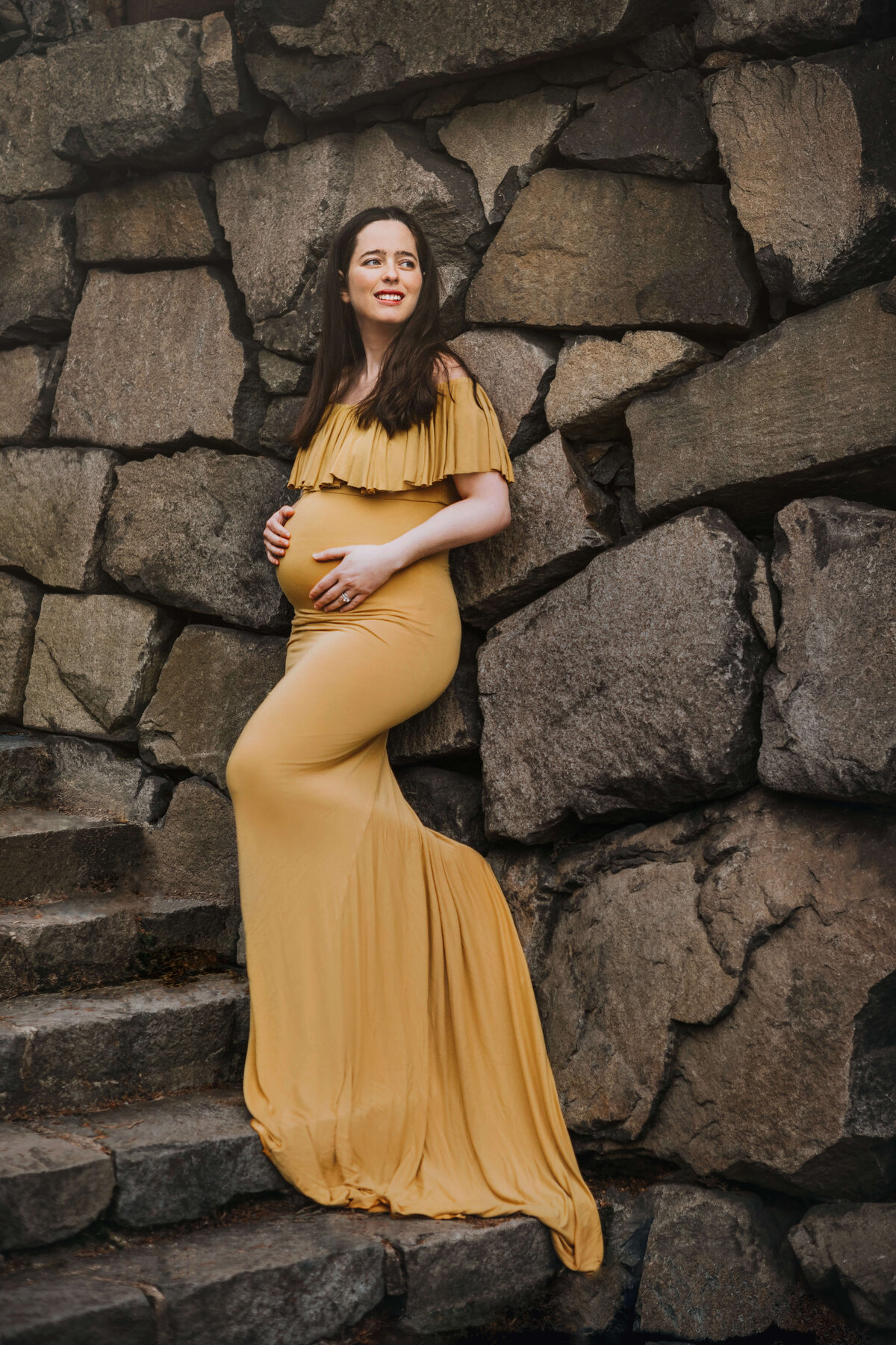 pregnant mother wearing a mustard maternity gown standing on stone steps leaning against a stone wall