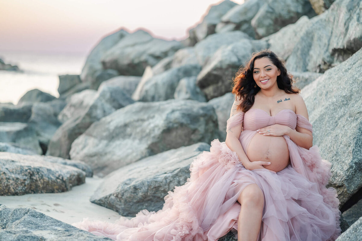 A pregnant woman sits on some rocks during her beach maternity session. She is wearing a pink tulle dress.