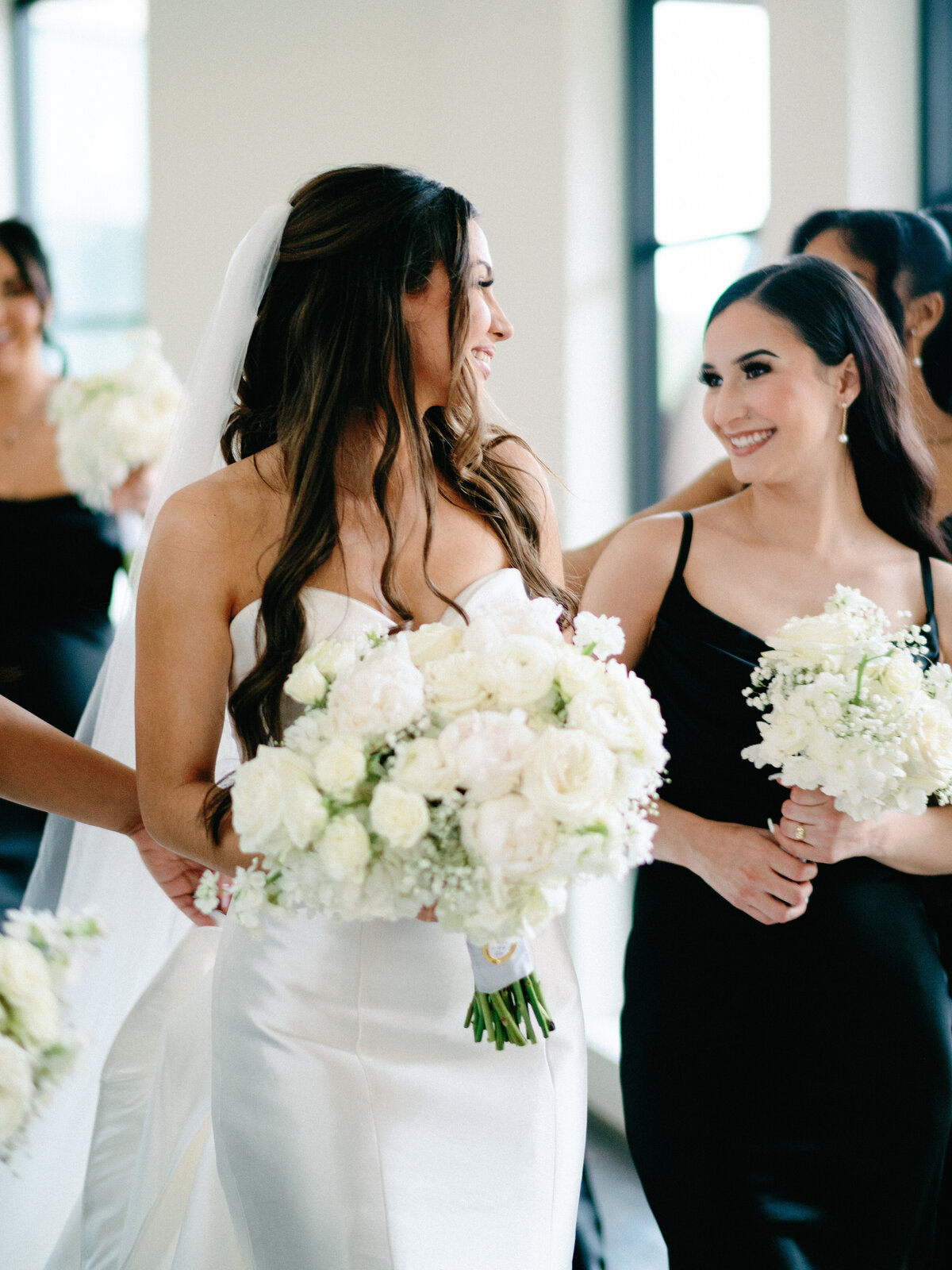 Modern bride walking and smiling at bridesmaids wearing black with white flowers