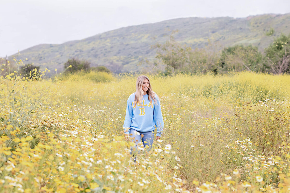 A person in a blue sweatshirt walks through a field of yellow and white wildflowers. Rolling hills are visible in the background under a cloudy sky.
