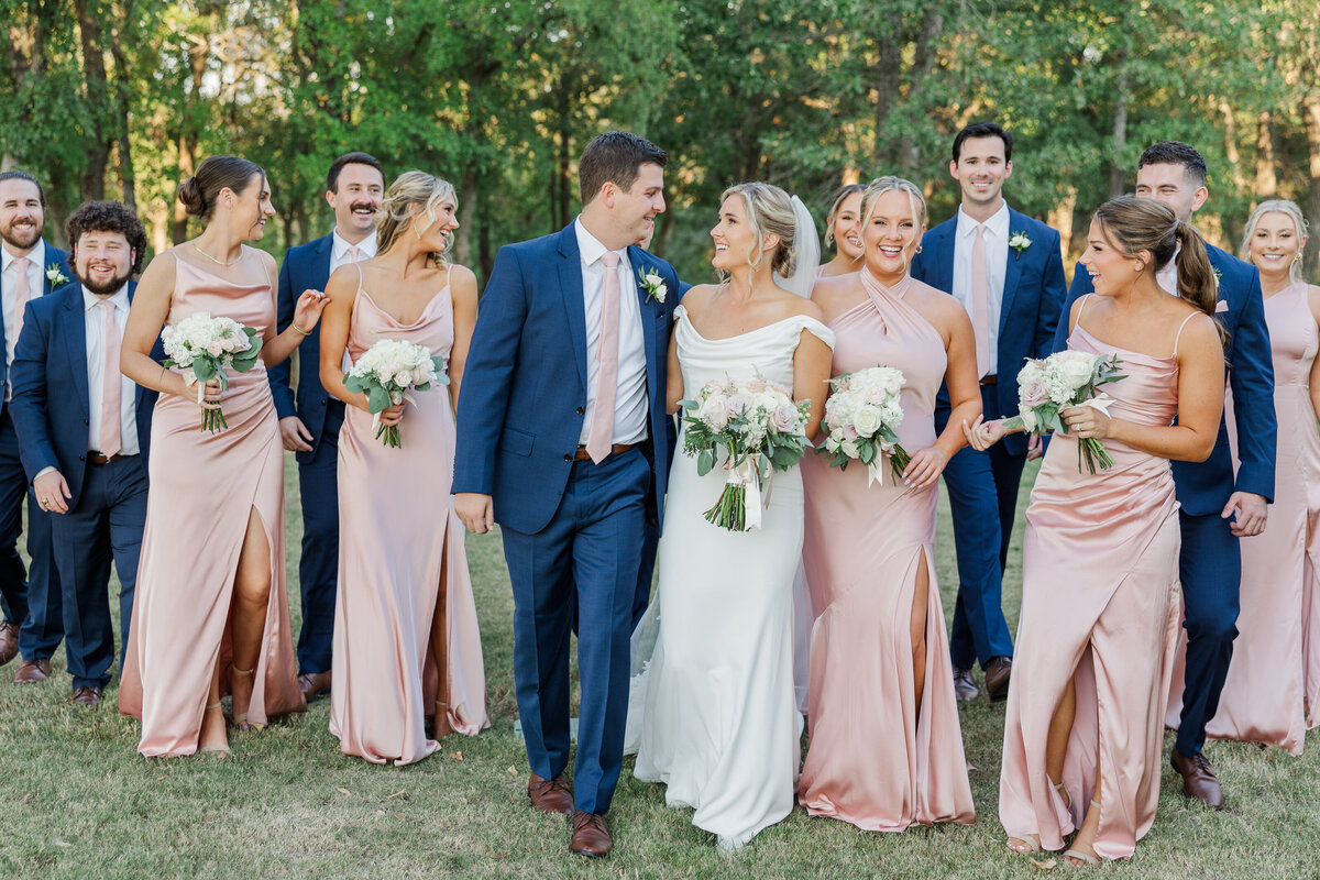 Bride and groom walk hand-in-hand with their joyful bridal party in soft blush and navy attire, sharing laughter in a candid moment outdoors. Captured in a timeless, light and airy wedding photography style near Plano, Coppell, and across the greater DFW area.