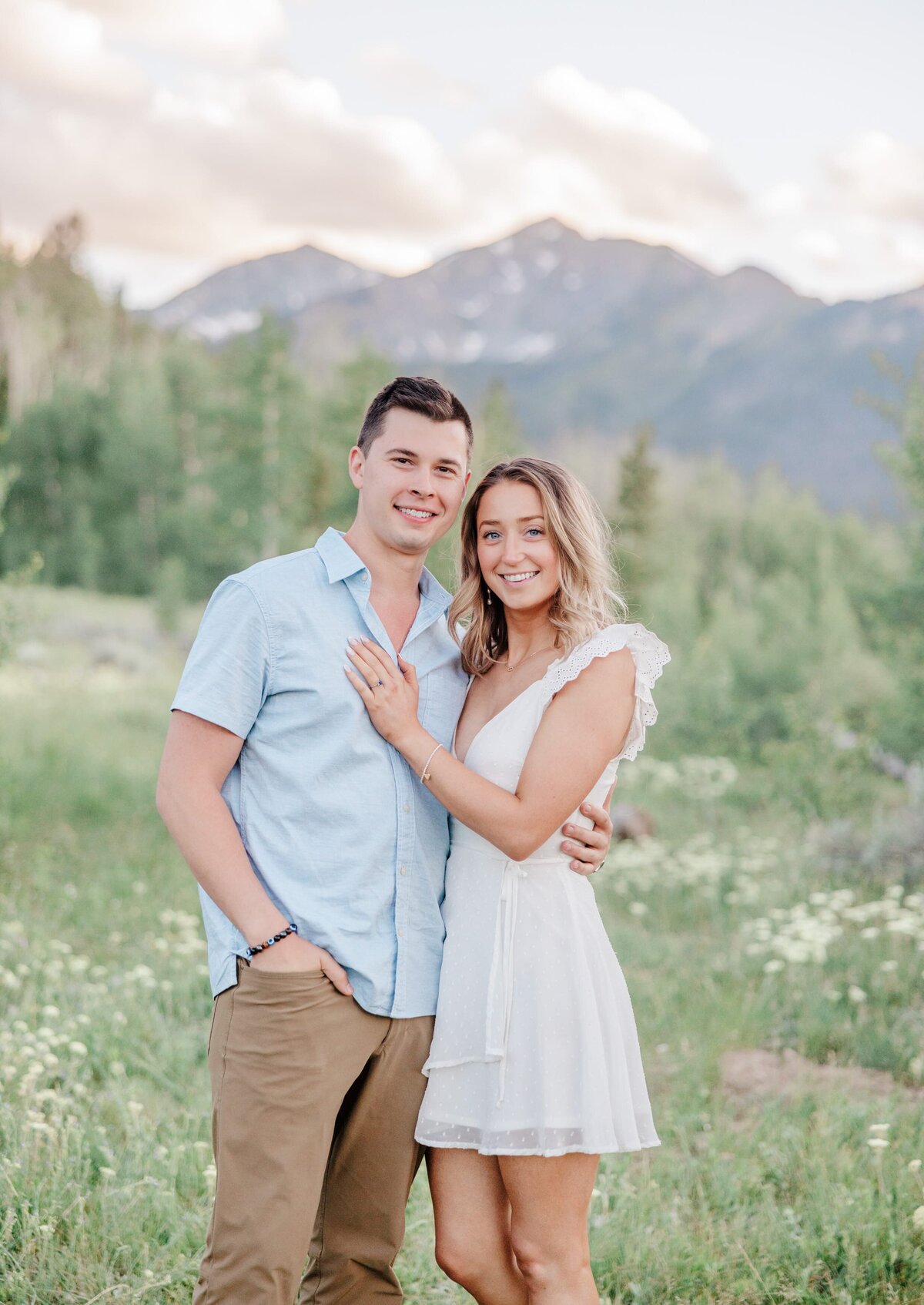 A woman in a white dress snuggles on her husband as they smile in a meadow filled with wildflowers