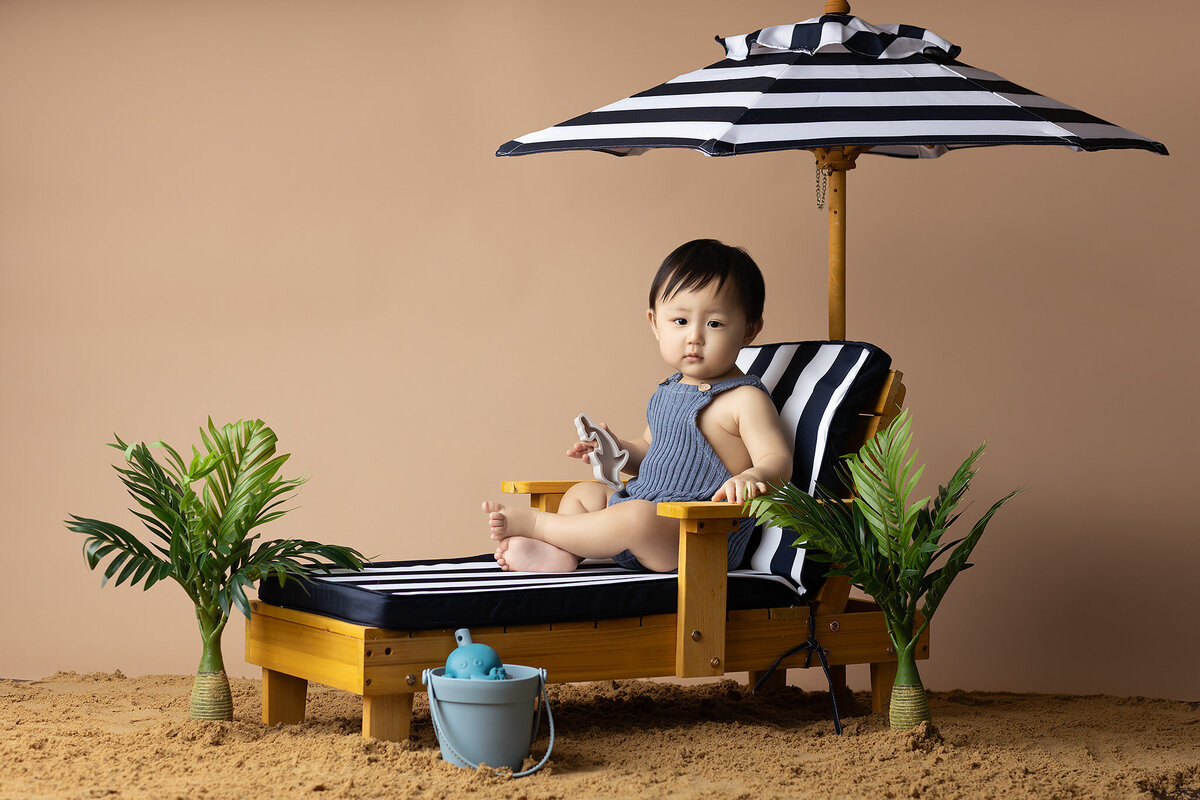 This adorable baby enjoys a beach-themed milestone photoshoot with a striped umbrella and tropical props. Capturing these moments through Dallas baby photography helps preserve the unique milestones of your little one's first year. Let Dallas baby photographers turn these moments into lasting memories with their artistic photography style.