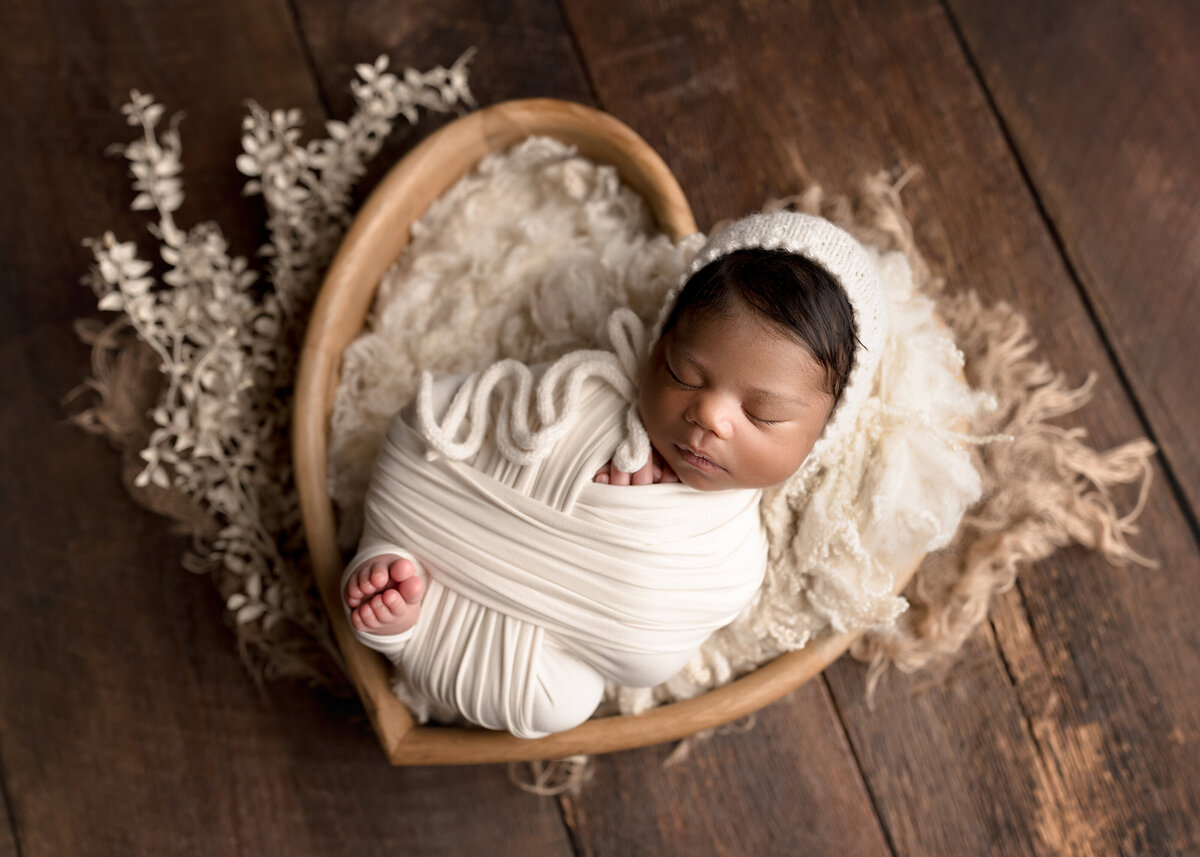 Black baby girl newborn photoshoot in West Palm Beach Florida. Baby is wrapped in a cream coloured knit swaddle with her toes peeking out and the bottom of her feet touching. Baby is wearing a matching knit bonnet. Baby is in a heart-shaped bowl with flowers beneath it.