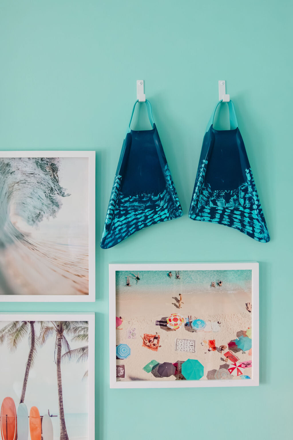 Surf-inspired kids’ bedroom wall featuring a pair of blue swim fins hung on a seafoam green wall above a gallery of coastal artwork. Framed beach prints include a wave crashing, surfboards with palm trees, and a colorful aerial photo of umbrellas on a tropical shoreline. Coastal decor meets playful, modern interior design with a bright, vacation-home vibe.