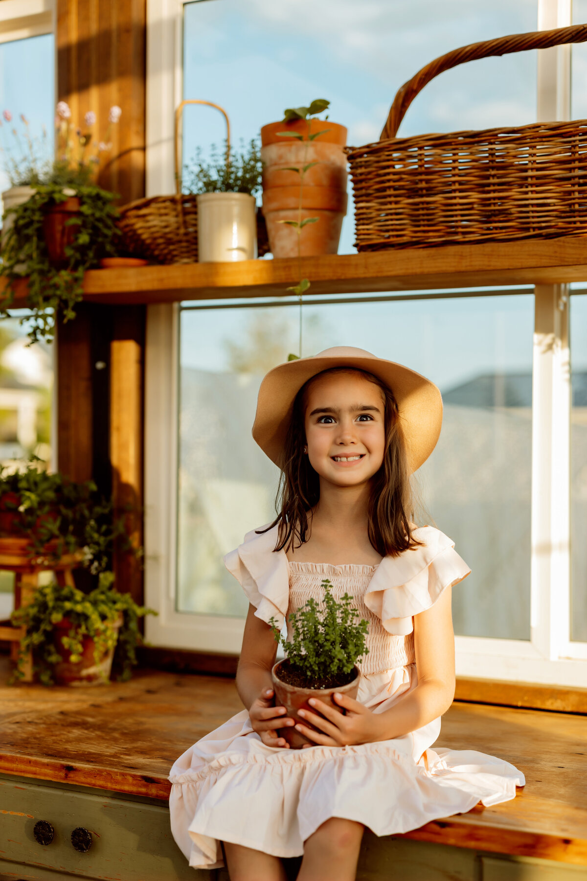 Young girl sits with her potted flower on a work bench in a green house for her family photo session in Denver