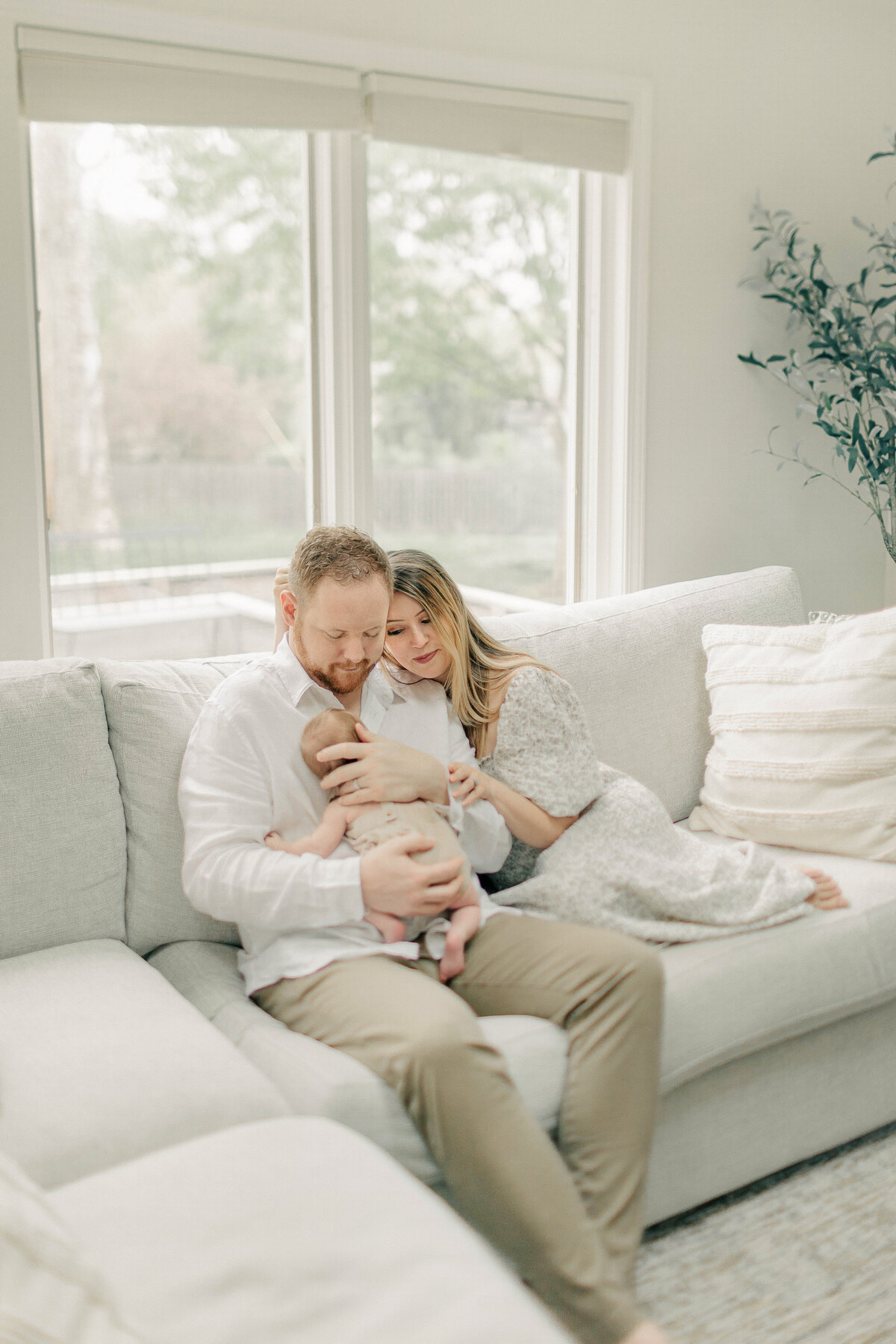 Mom and dad sitting on a light-colored couch with their newborn cuddled on dad's chest during a lifestyle newborn photo session at their Kansas City home.