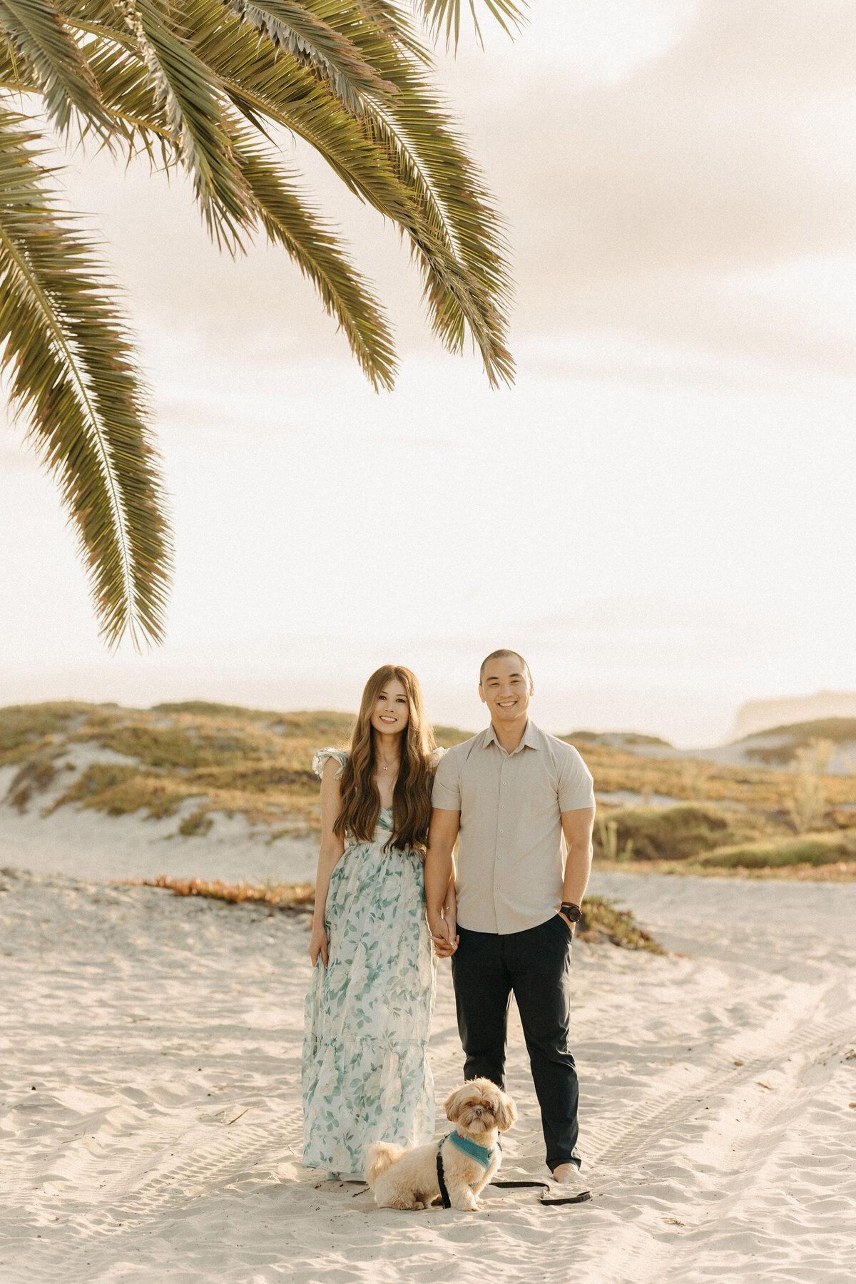 Couple standing on Coronado Beach at sunset with their small dog, holding hands under palm trees.