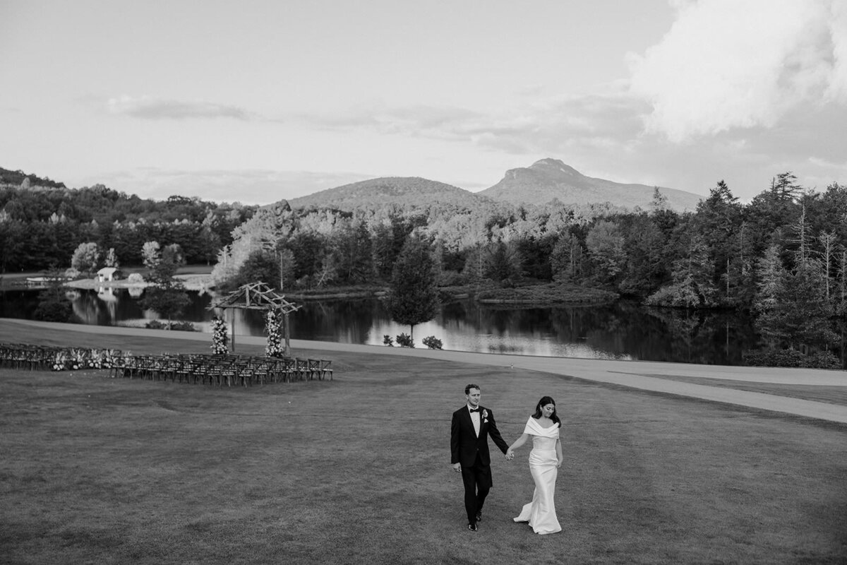 Bride and groom walk hand in hand across North Carolina field with wedding ceremony space in the background