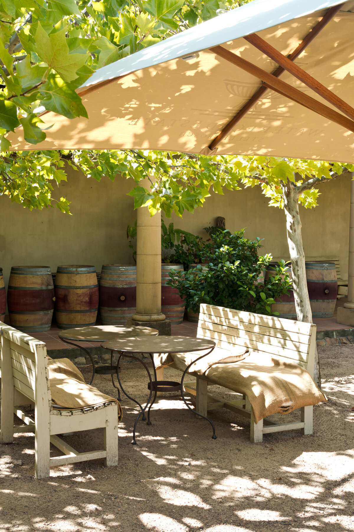 Outdoor seating area with a rustic table and benches beneath a large beige umbrella. Wooden barrels line the wall in the background, and leafy trees cast shadows on the sandy ground, creating a peaceful, shaded atmosphere.