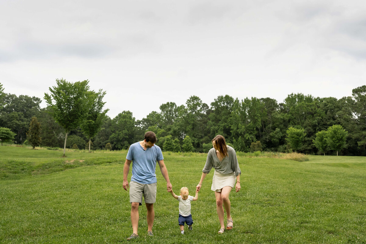 Parents holding hand of 1 year old boy while walking across grassy field.