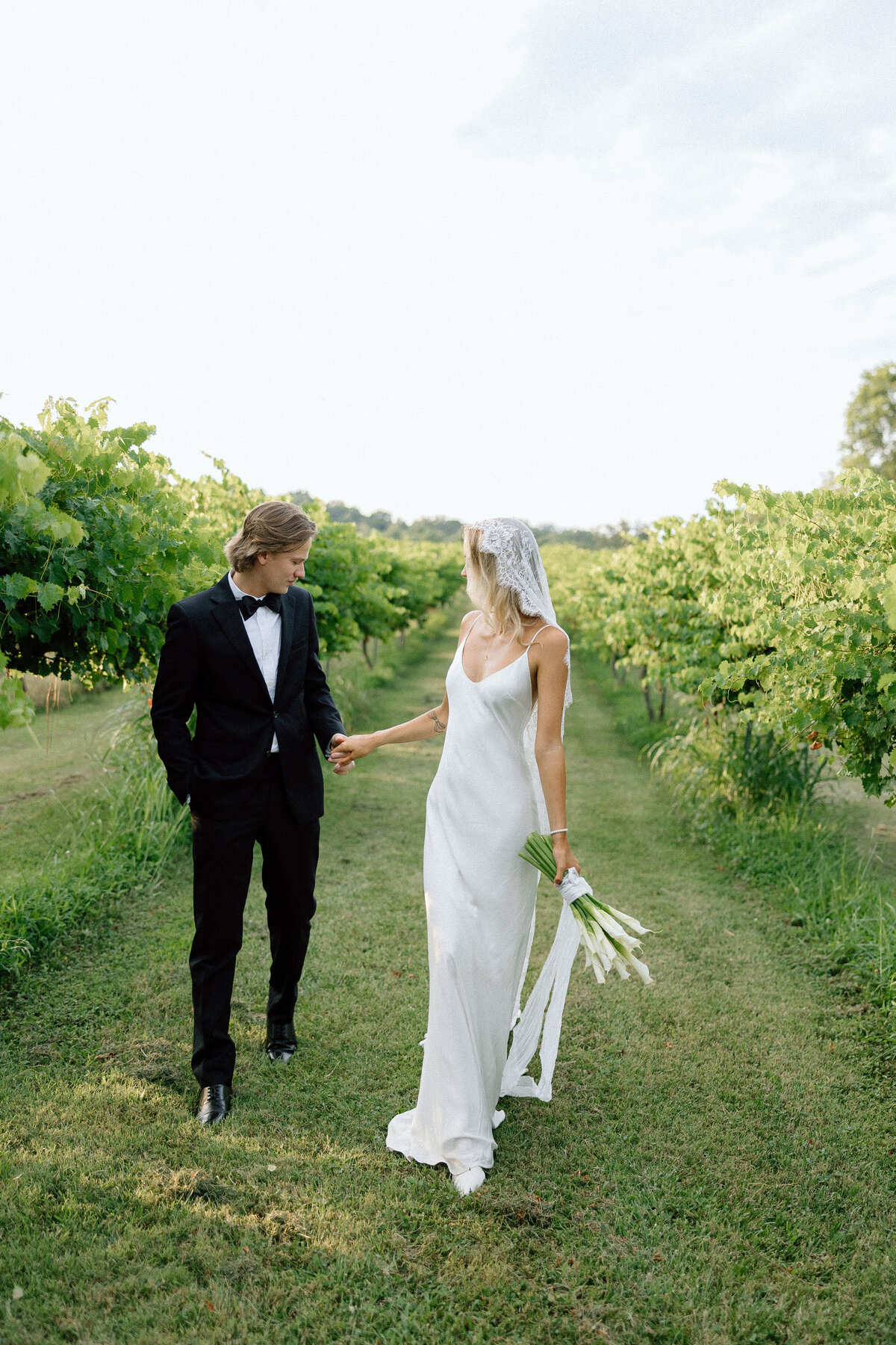 A couple holds hands as they walk together through the vineyard, looking backwards. 