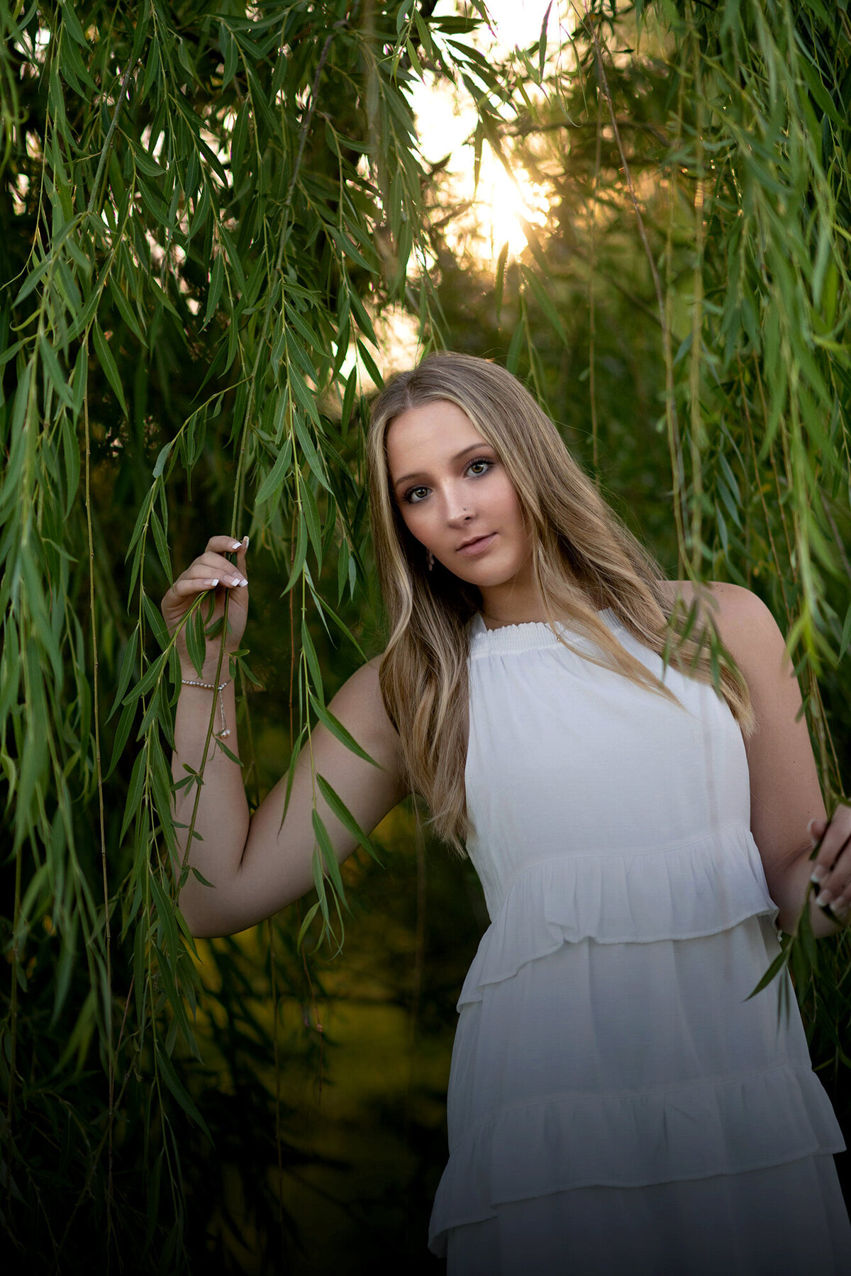 A high school senior in a white dress plays in the branches of a willow tree