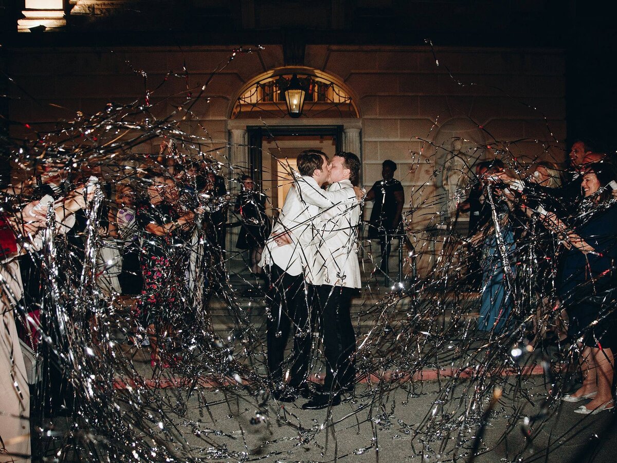 LGBTQiA wedding at Cheekwood Botanical Gardens with a silver streamer send off for two grooms wearing tuxedos with black pants and white jackets. The grooms are kissing as their guests cheer in front of the Cheek mansion.