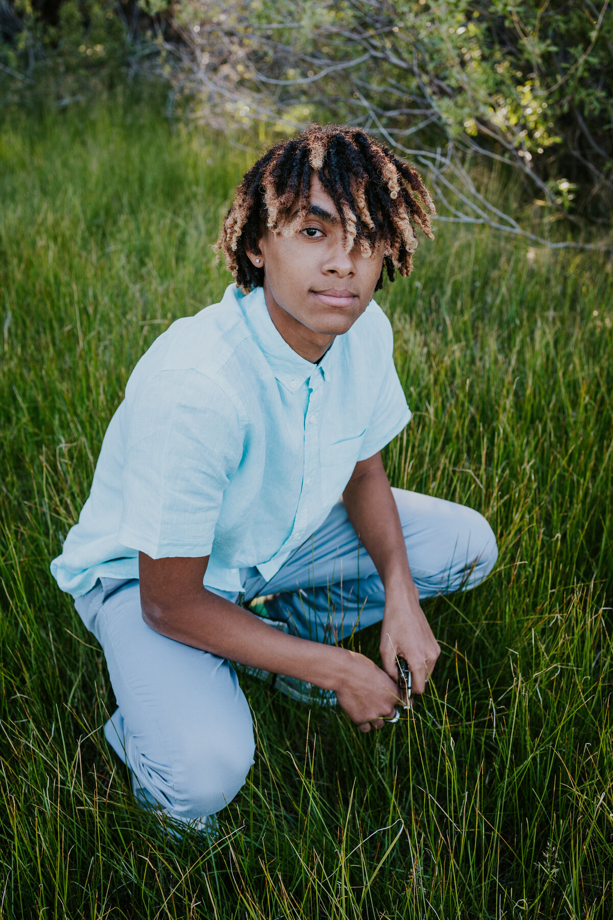 Young man squats down in grassy meadow while looking up at camera.