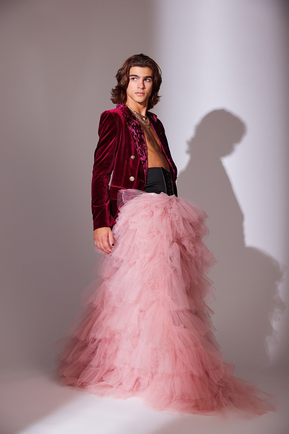 Male model in a dramatic pink tulle skirt and red velvet blazer, standing in studio lighting with shadow play on the wall—editorial-style senior portrait with fashion-forward styling