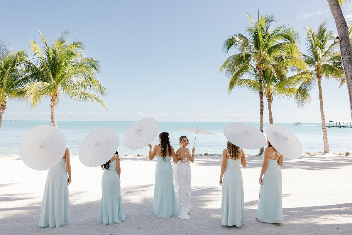 Ashley and her bridesmaids, with white parasols, walk hand in hand along the shoreline, surrounded by palm trees.