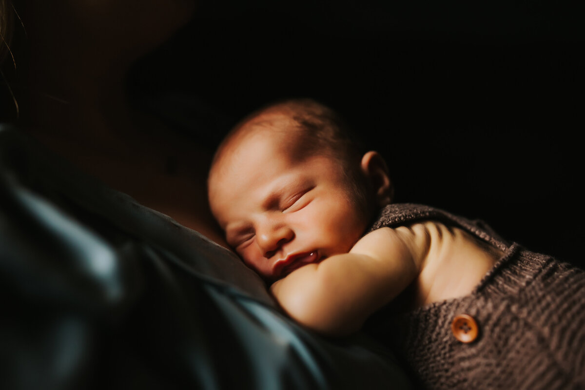 sleepy newborn baby on his mothers chest wearing a knit overall jumpsuit with window light hitting his face
