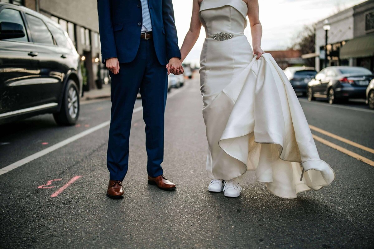 A bride in a white wedding dress and sneakers holds up her gown while standing on a city street, holding hands with a groom in a blue suit. Their faces are not visible. Cars are parked along the street.